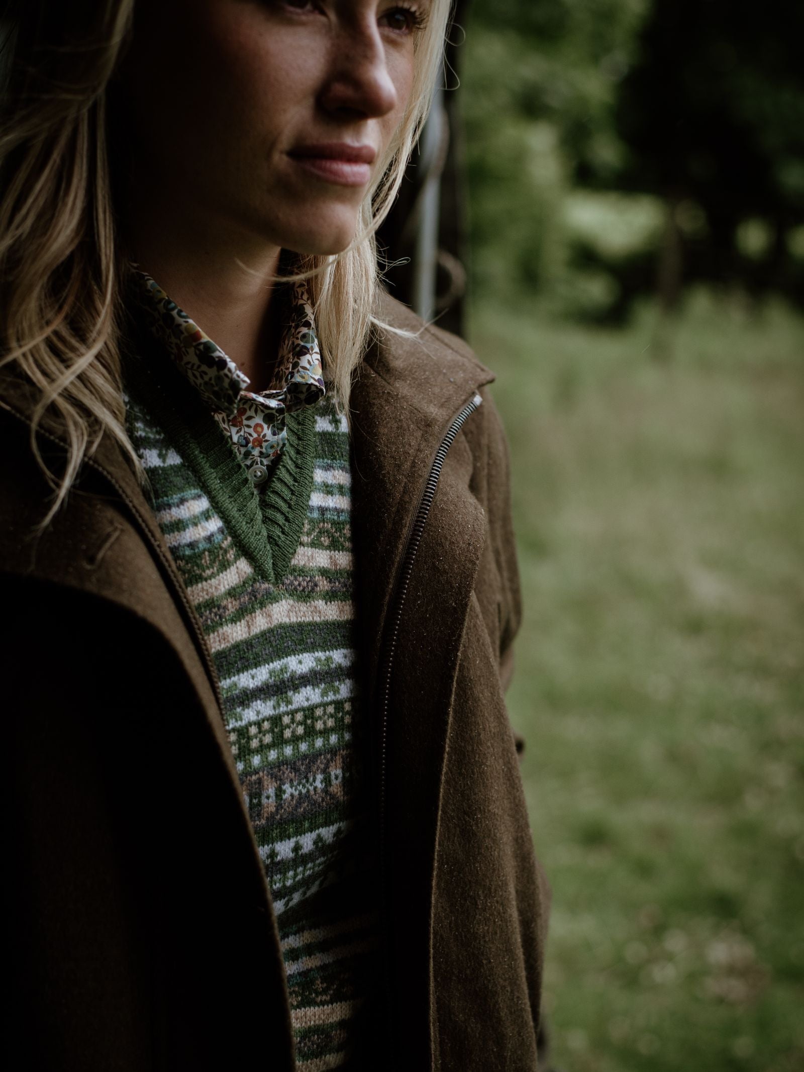A woman with blonde hair wears the Campbell's of Beauly Cashmere V-Neck Fairisle Slipover over a floral collared shirt and brown coat, standing in a grassy, wooded area. The photo shows her from the shoulders up.