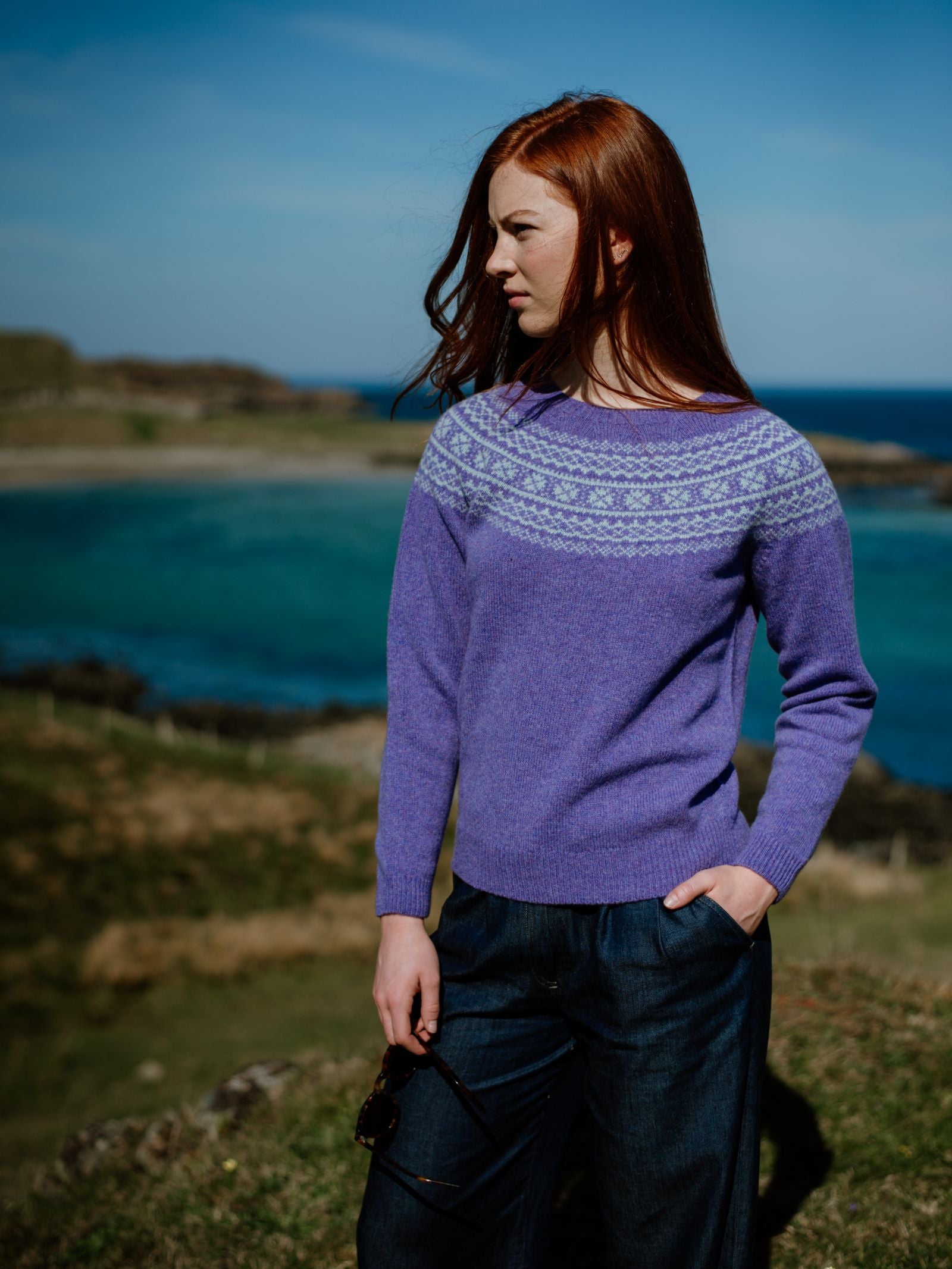 A woman with long red hair, wearing Campbells of Beauly Two-Colour Fairisle Yoke Jumper and dark pants, stands outdoors with one hand in her pocket. Grassy hills, rocky terrain, and a calm blue sea stretch out behind her under a clear sky.