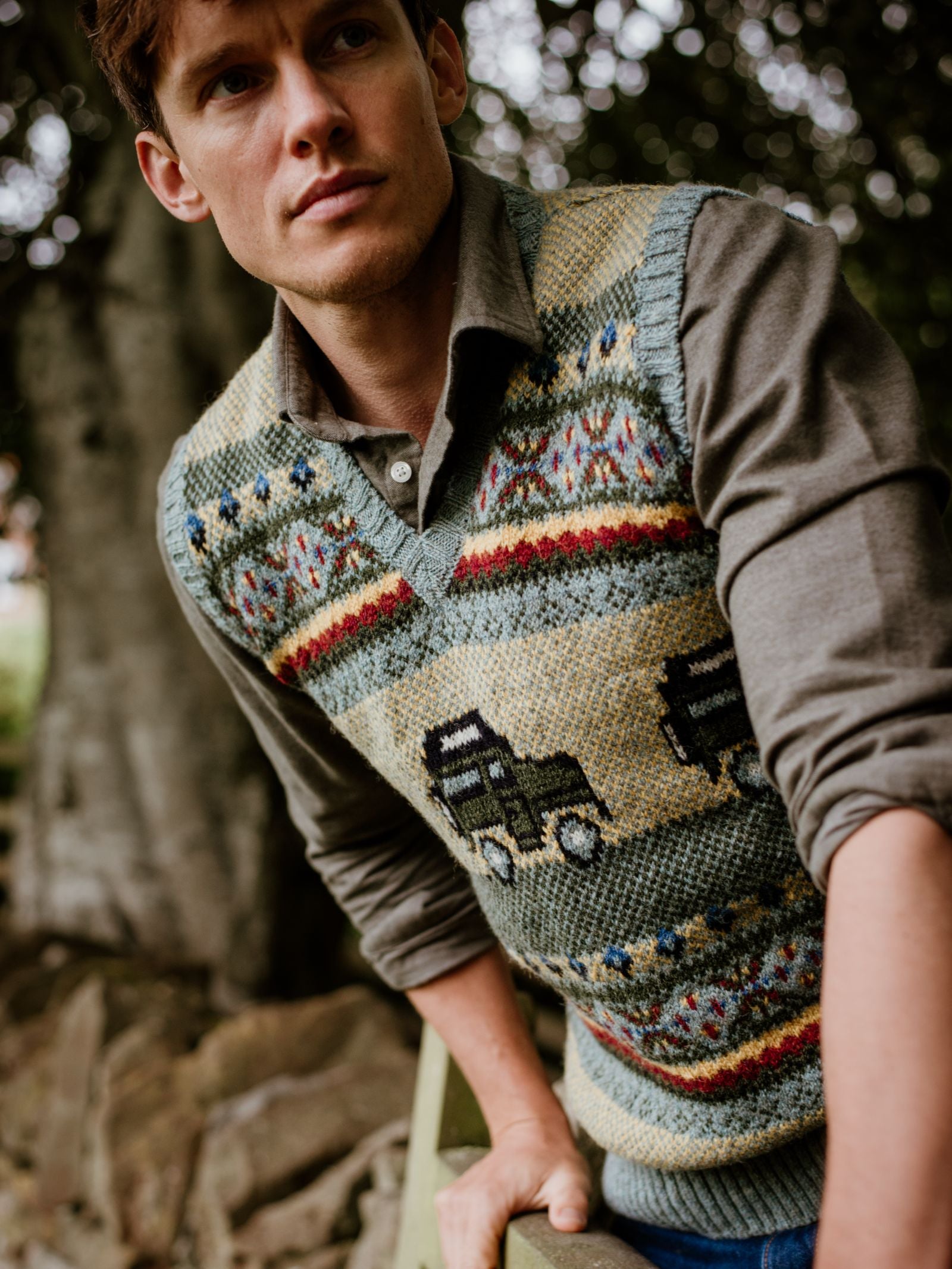 A man in the Campbells of Beauly Land Rover Defender Fairisle Slipover, featuring vehicle and decorative designs, leans on a wooden fence outdoors with trees and rocks in the background.