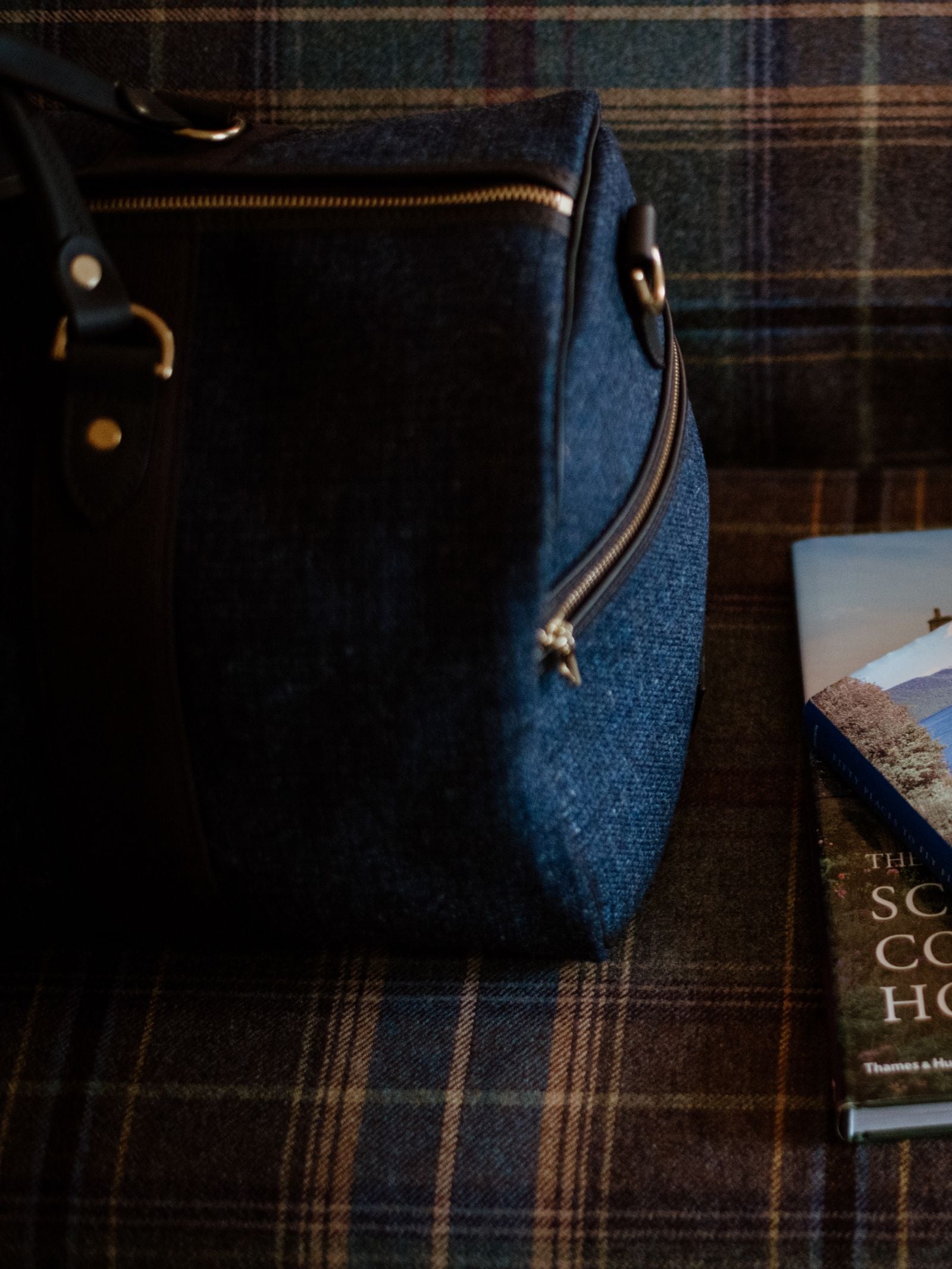 A Campbell's of Beauly Hand Luggage Holdall with black straps sits on a plaid surface beside a scenic-covered book, making it the ideal travel companion.