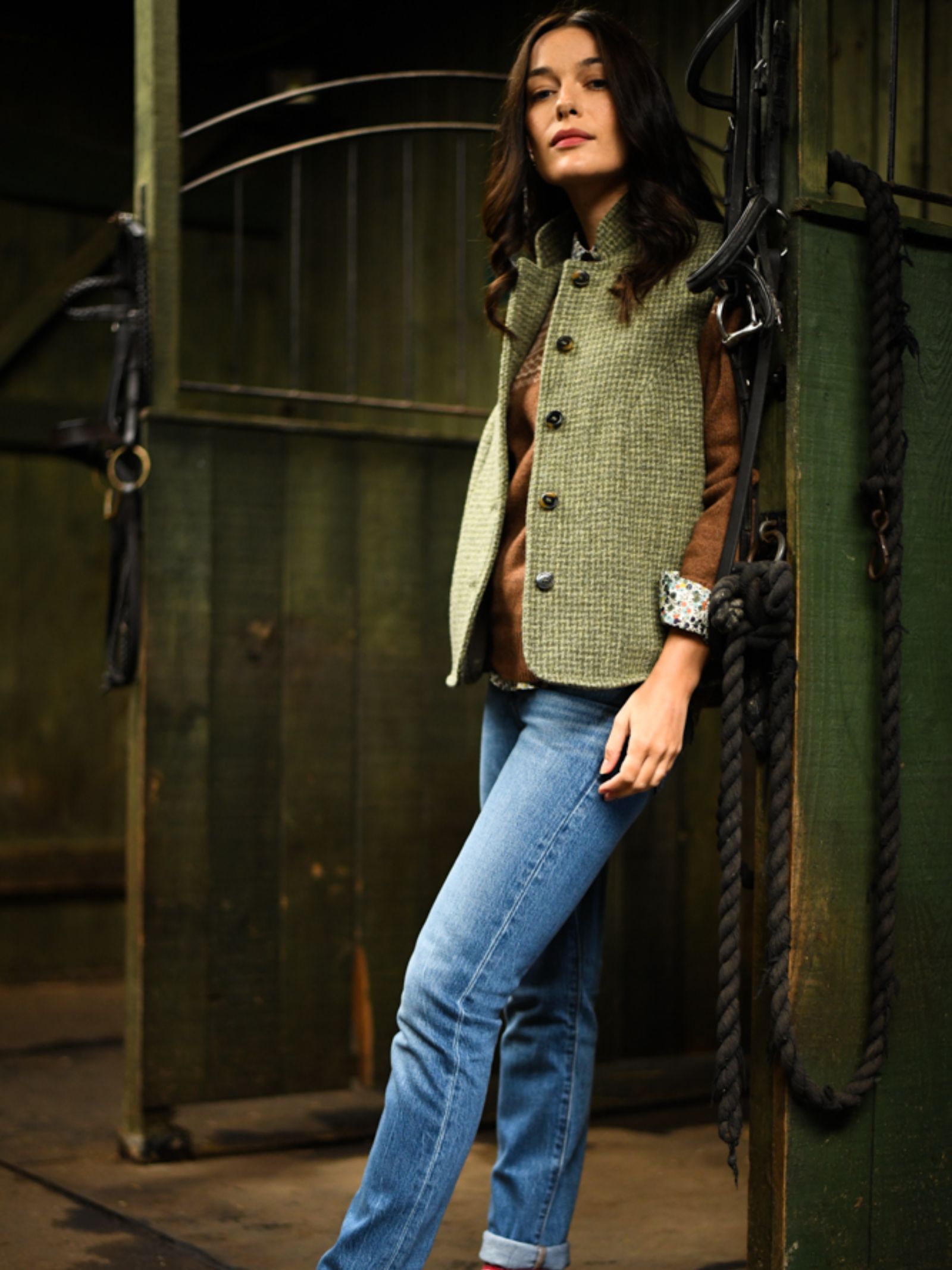 A woman wearing the Harris Tweed Houndstooth Gilet by Campbell’s of Beauly, styled with a brown sweater and blue jeans, leans against a stable door framed by dark green wood walls and black ropes.