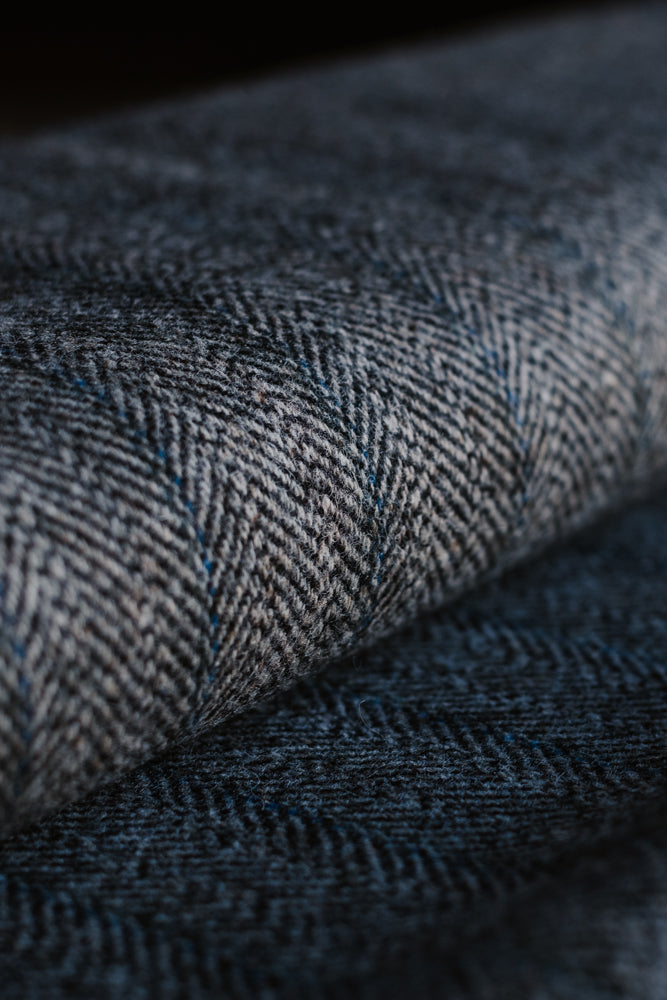 Close-up of a roll of Campbell’s of Beauly House Tweed 95053, featuring a dark gray and blue herringbone pattern with detailed texture and diagonal woven lines—an ideal jacketing cloth that evokes the classic look of traditional herringbone tweed.