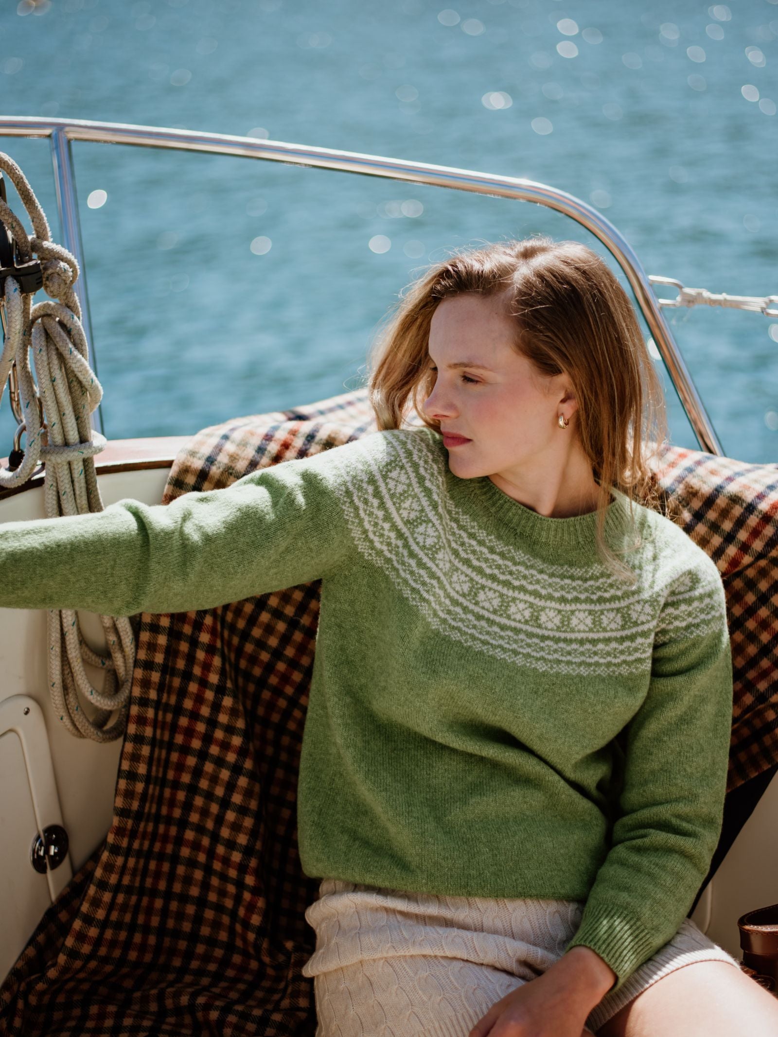 A woman with light brown hair sits on a boat, wearing Campbell’s of Beauly’s Two-Colour Fairisle Yoke Jumper with a cream skirt. Calmly looking left, she is framed by rope, a checkered blanket, and sparkling blue water.