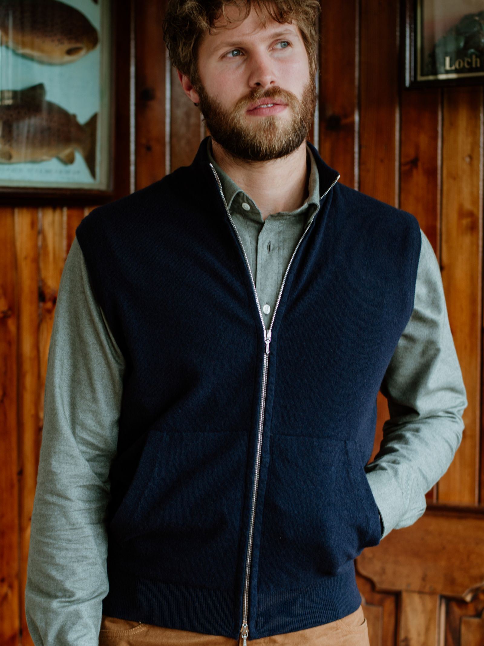 A man with curly brown hair and a beard wears a light green Campbells of Beauly Twill Shirt in classic fit, layered under a navy zip-up vest with brown pants. Indoors, he stands by wood-paneled walls and framed fish art, gazing slightly to the side.
