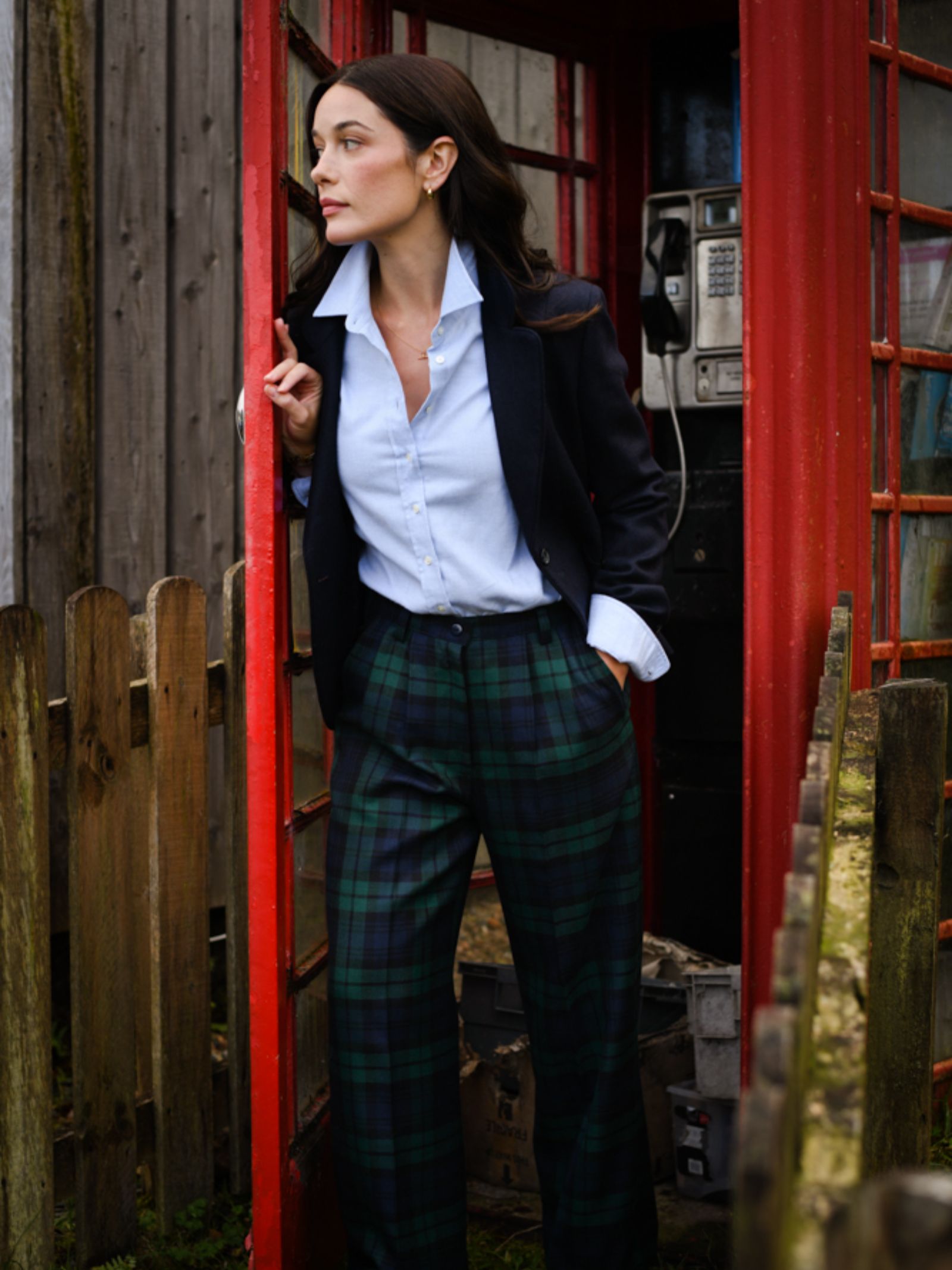 A woman in a navy blazer, white shirt, and green plaid pants stands by a classic red British phone booth, looking to the side. She completes her look with the gold herringbone House Jacket from Campbell's of Beauly. A wooden fence is in the background.