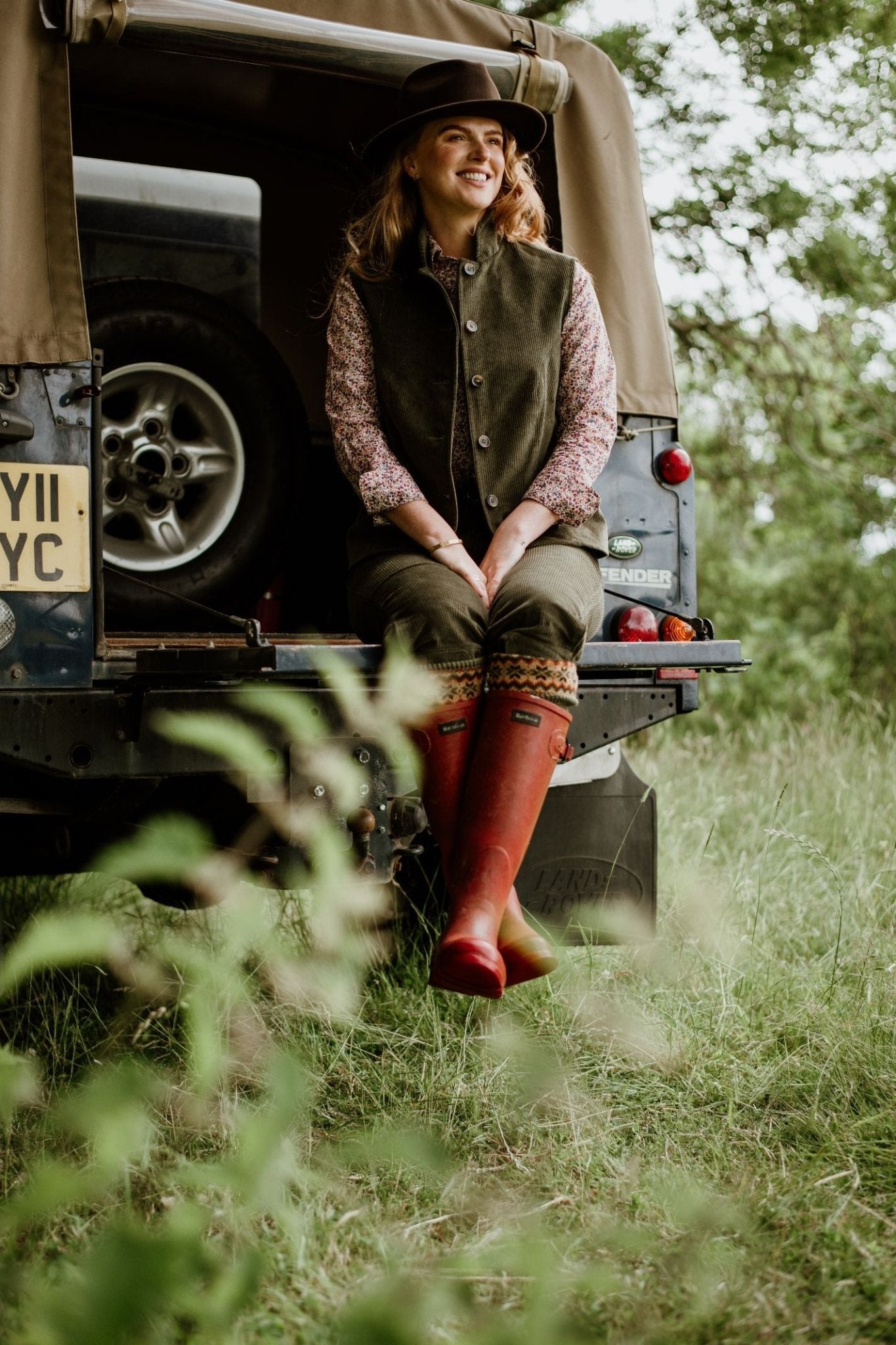 A woman radiates rustic charm in a Campbell's of Beauly Corduroy Gilet and floral shirt, paired with red rain boots, as she sits smiling on an off-road vehicle in the grass.