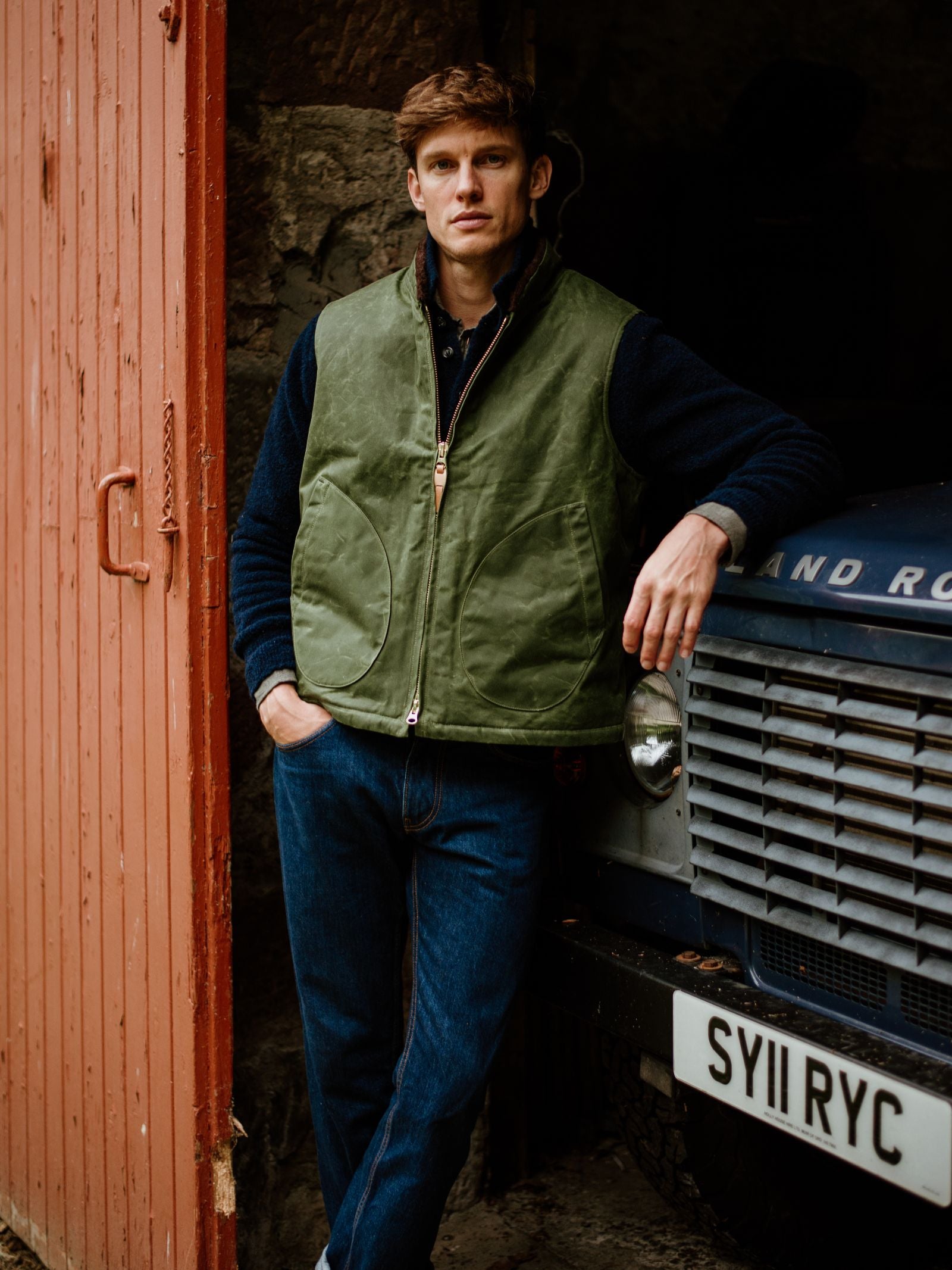 A man in a green Wax Nehru Gilet by Campbell's of Beauly and blue jeans stands with one hand in his pocket and the other on a vintage blue Land Rover inside a rustic building with a red wooden door.