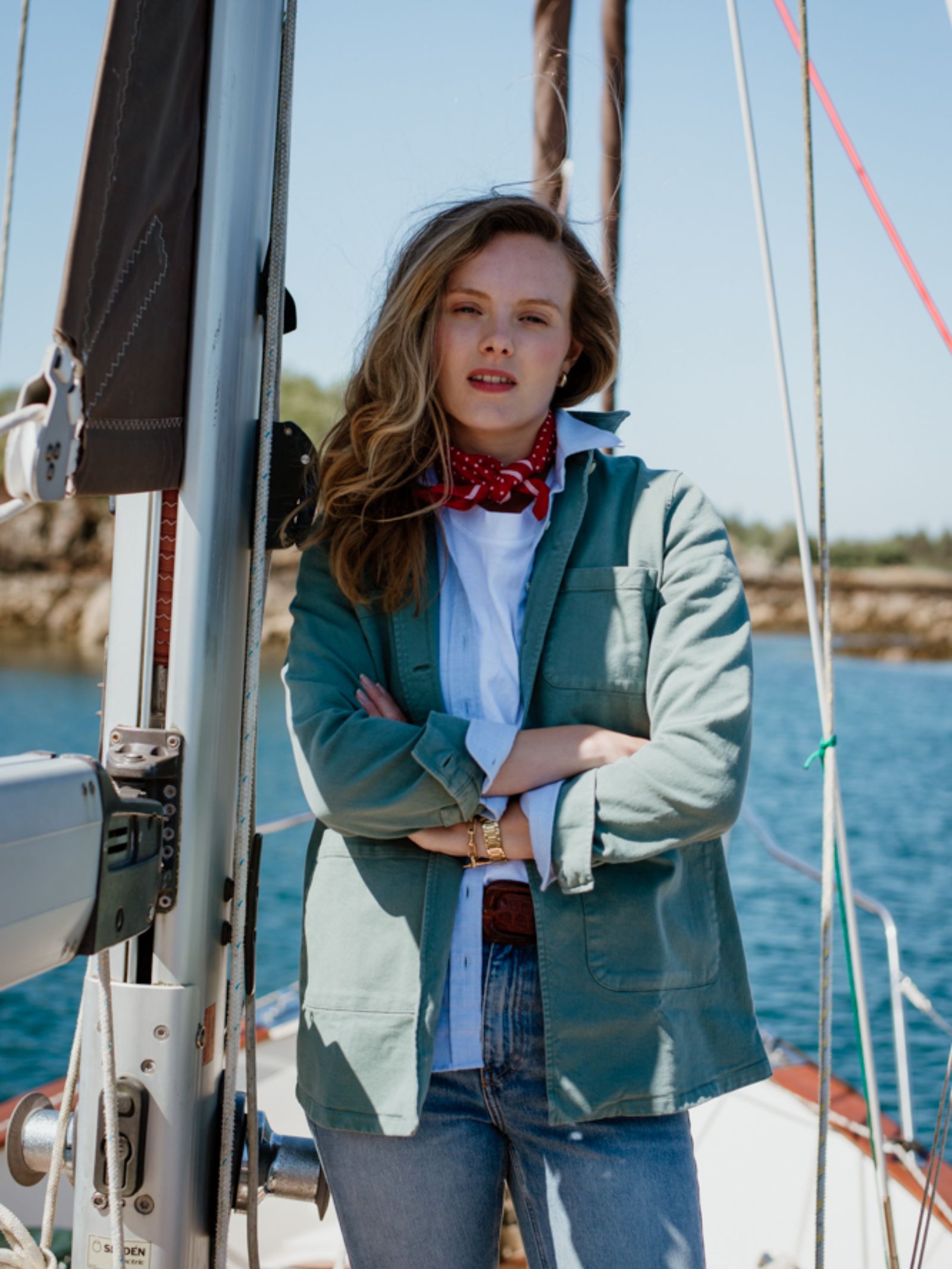 A woman stands confidently on a sailboat, wearing the Campbells of Beauly Summer Chore Jacket over a white shirt and jeans. She accessorizes with a red patterned scarf and brown belt, as sunlight glimmers on the water behind her.