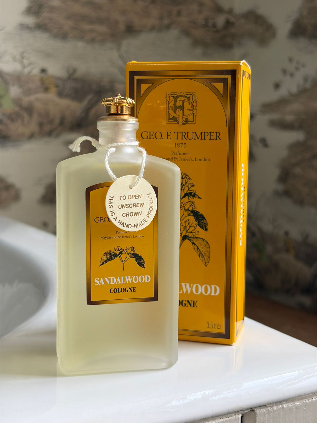 A frosted glass bottle of Geo. F. Trumper Sandalwood Cologne 100ml sits beside its yellow box on a bathroom counter against nature-themed wallpaper.