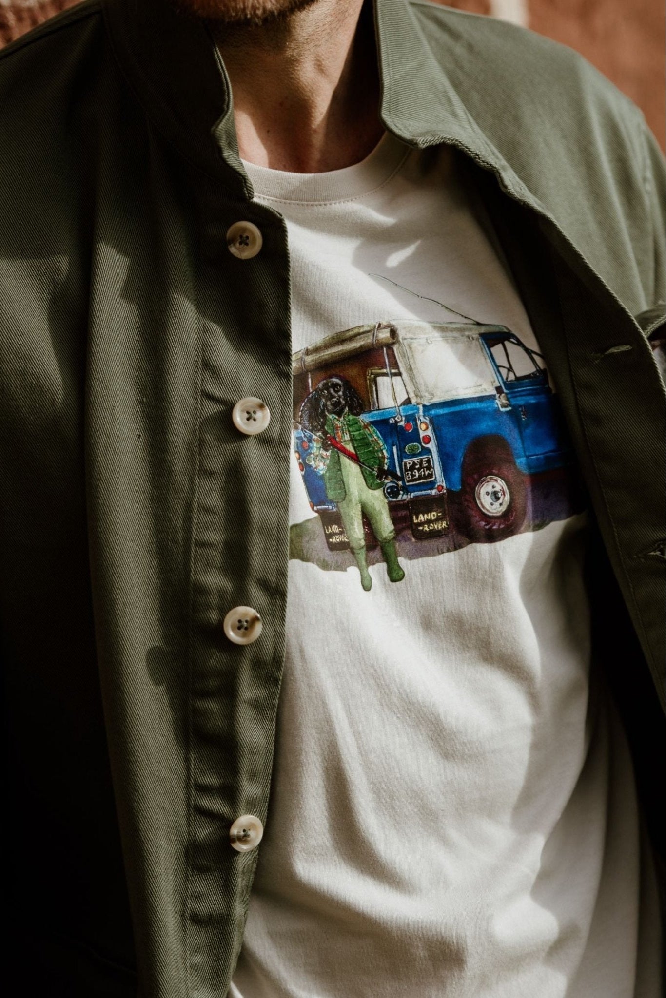 A person wearing an olive green jacket over a Campbells of Beauly Land Rover Cocker T-Shirt, which is white cotton and features a graphic of a blue Land Rover with a person standing in front. Only the upper torso is visible.