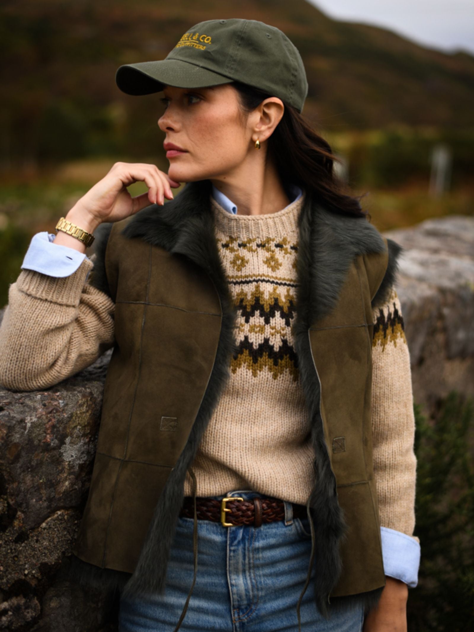 A woman in a green cap, patterned sweater, and jeans stands by a stone wall, wearing the Sheepskin Reversible Gilet by Campbell's of Beauly. She gazes to the side with greenery and hills in the background.