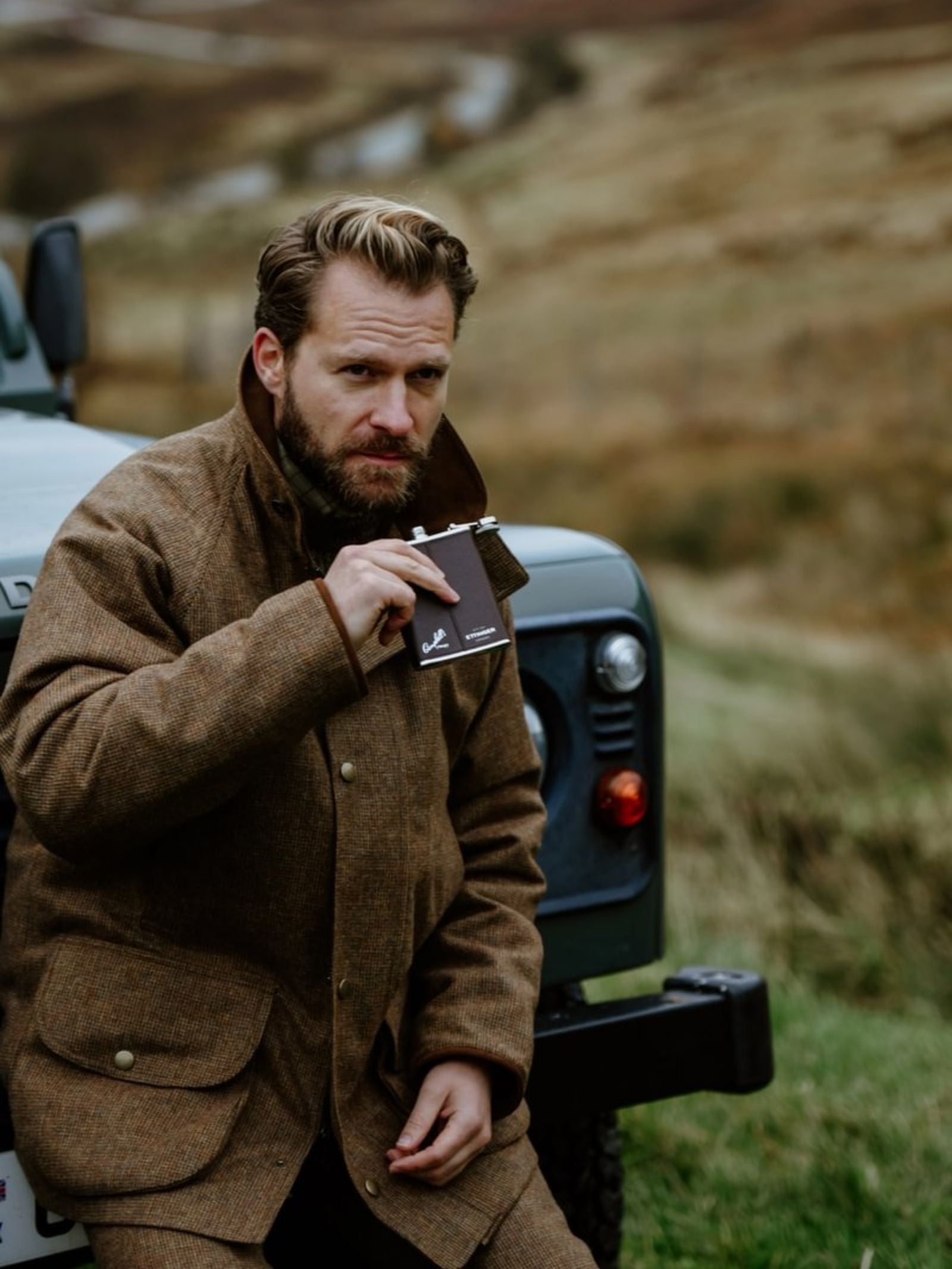 A bearded man in a brown tweed jacket sits on a green vehicle in the countryside, thoughtfully holding the 6oz Chocolate Hip Flask by Campbell's of Beauly x Ettinger—capturing timeless British style.