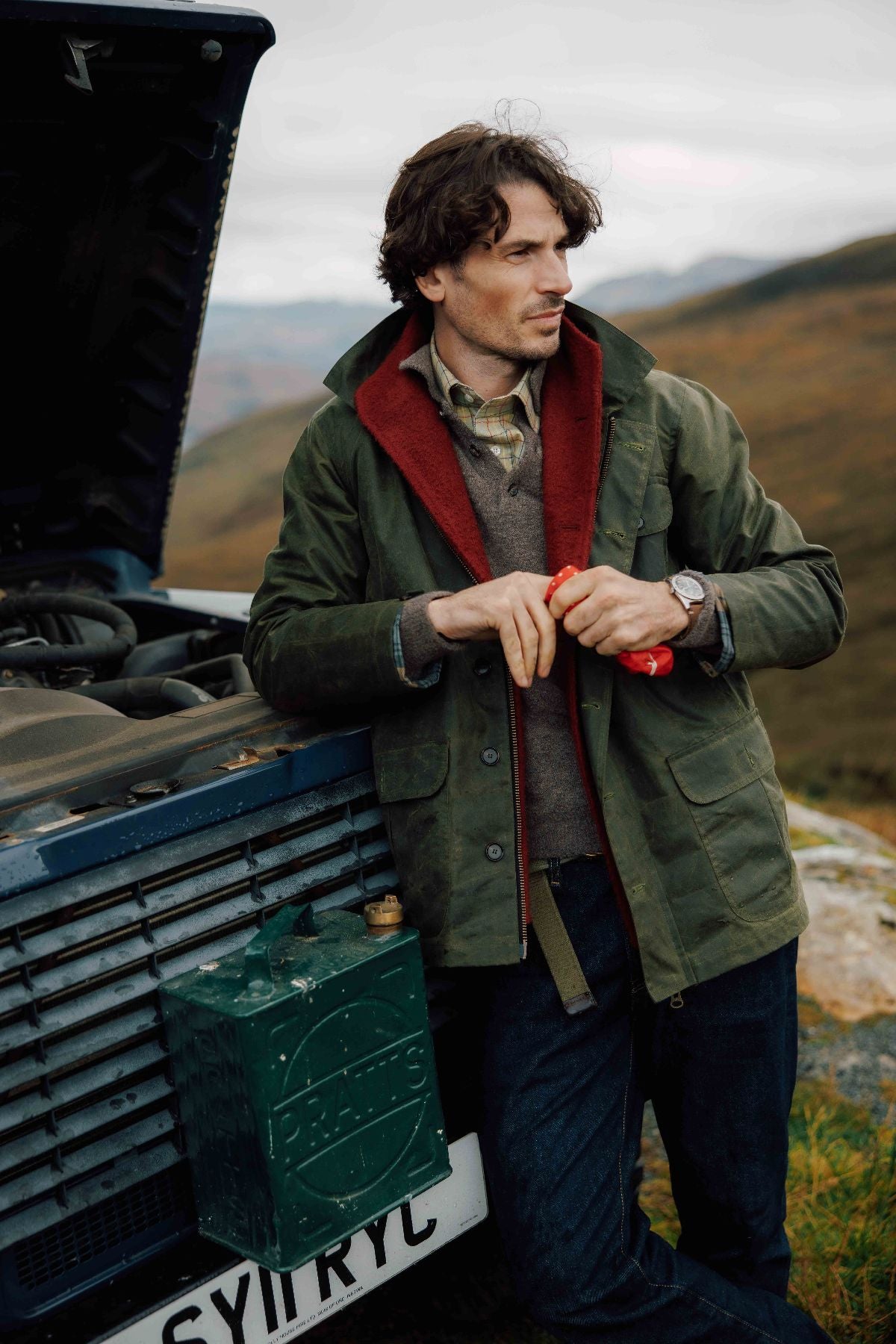A man in the Garve Jacket by Campbell's of Beauly and a red scarf leans against a blue vehicle with its hood open on a rural road, holding a red item and gazing into the distance with hills in the background.
