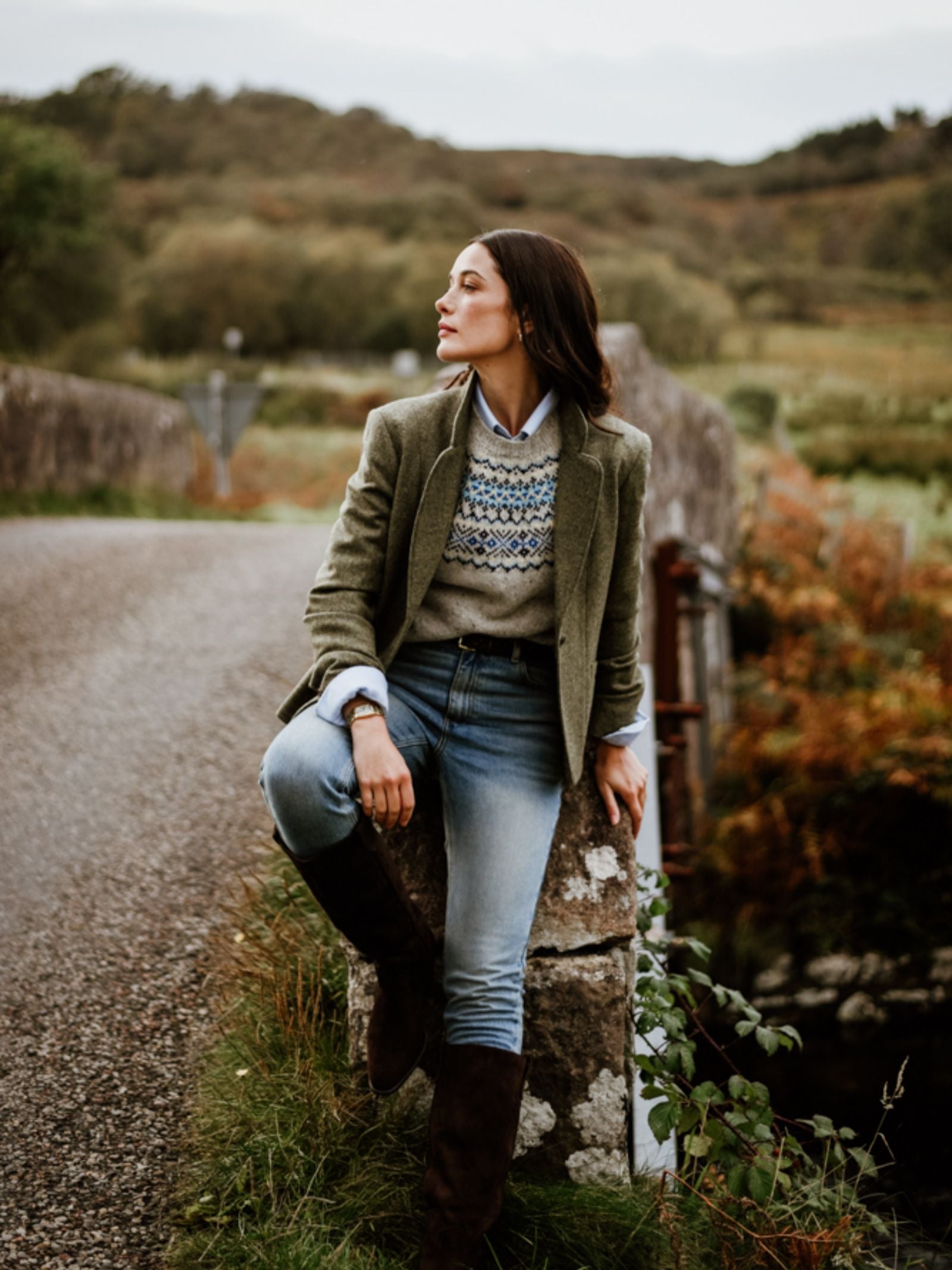A woman wearing the Campbell’s of Beauly House Jacket sits on a stone post by a country road, surrounded by lush green and autumnal scenery. She gazes to the side, appearing relaxed and thoughtful in the classic tweed look.