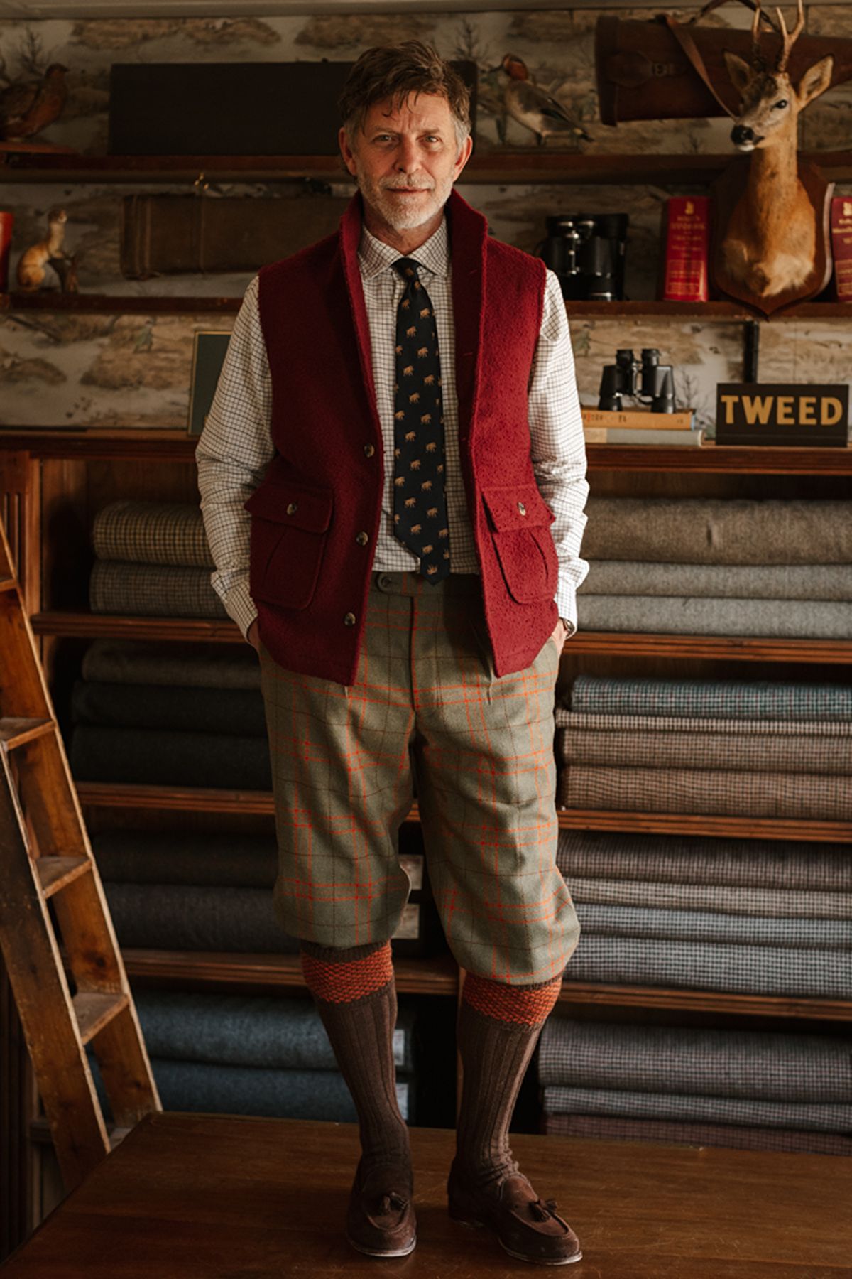 A man wearing a red waistcoat, checked shirt, elephant-patterned tie, and grey checked breeches standing in front of shelves filled with rolls of tweed fabric.