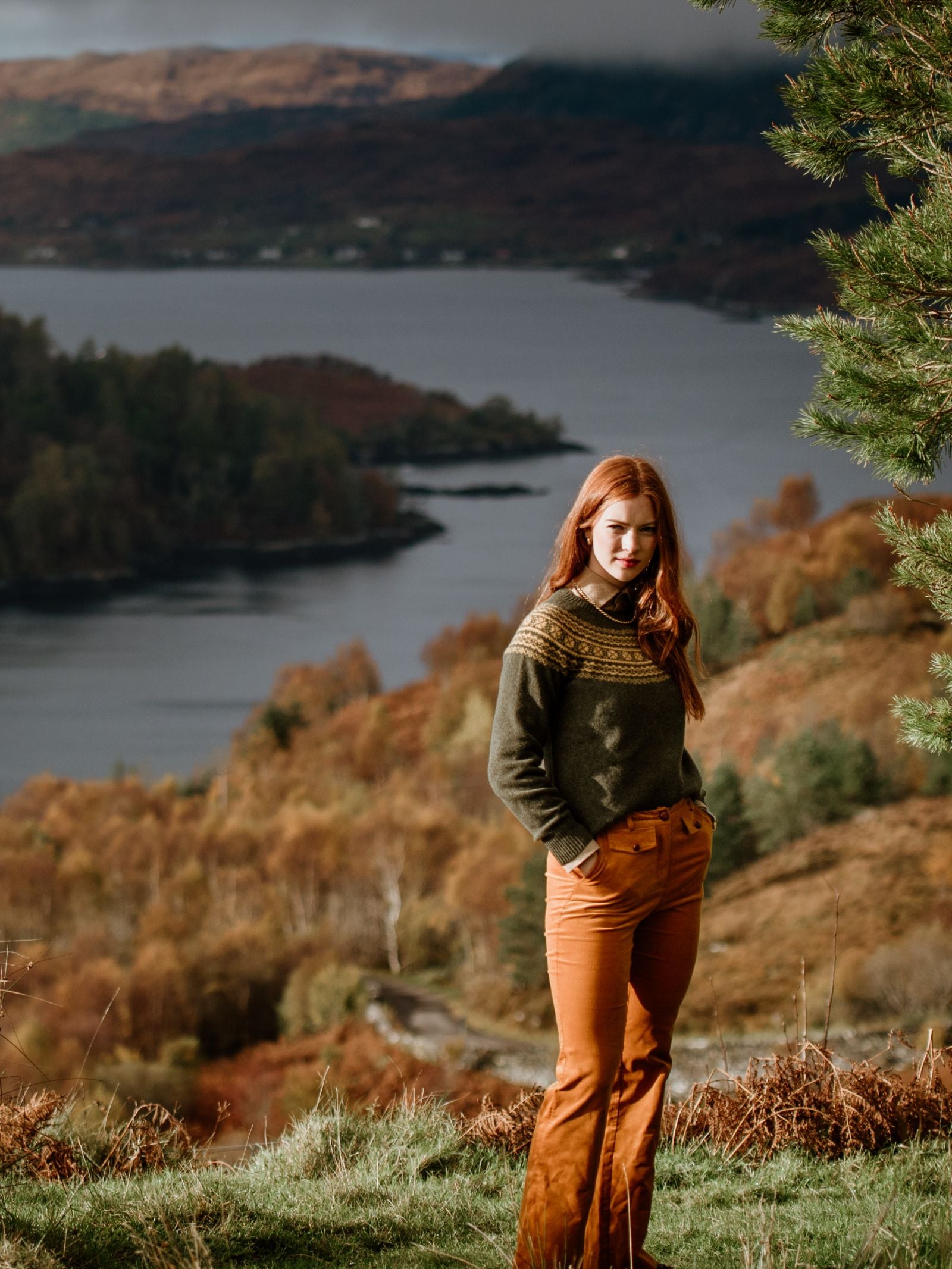 A woman with long red hair stands on grass, wearing an olive-green Campbells of Beauly Two-Colour Fairisle Yoke Jumper and rust-colored pants. Behind her are autumn trees and a lake set among hills under a cloudy sky.