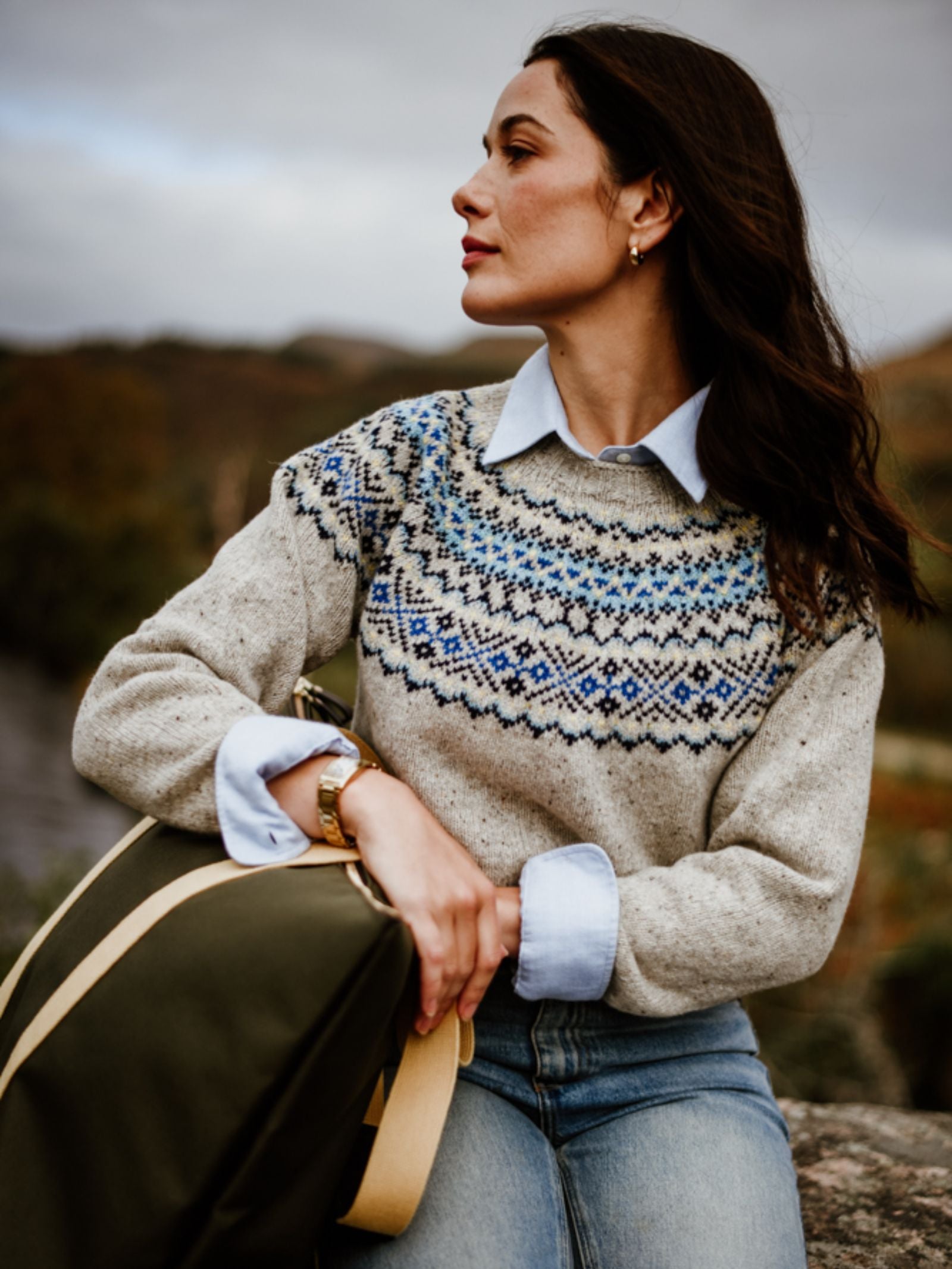A woman with long brown hair sits outdoors, looking to the side. She wears the Donegal Lambswool Fairisle Crew by Campbell's of Beauly over a collared shirt, jeans, gold hoop earrings, and holds a green bag. The background is blurred and natural.