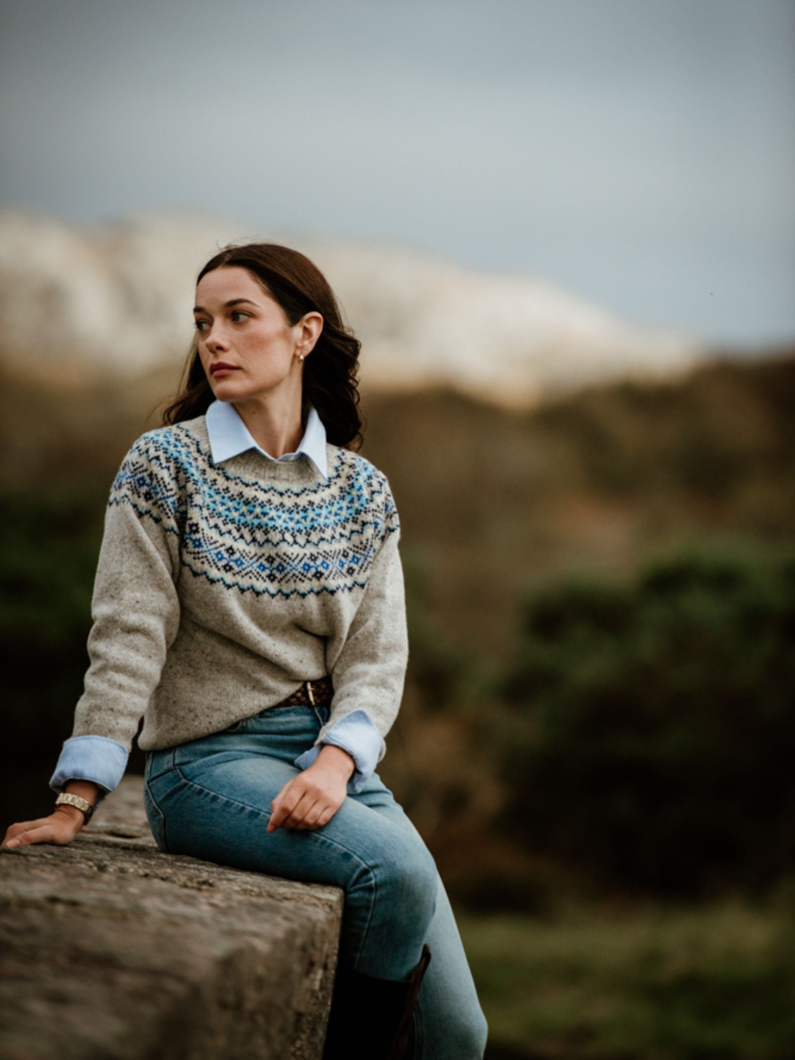 A woman wearing the Campbell's of Beauly Donegal Lambswool Fairisle Crew Jumper and jeans sits on a stone wall outdoors, gazing to the side, with blurred trees and hills under a cloudy sky in the background.