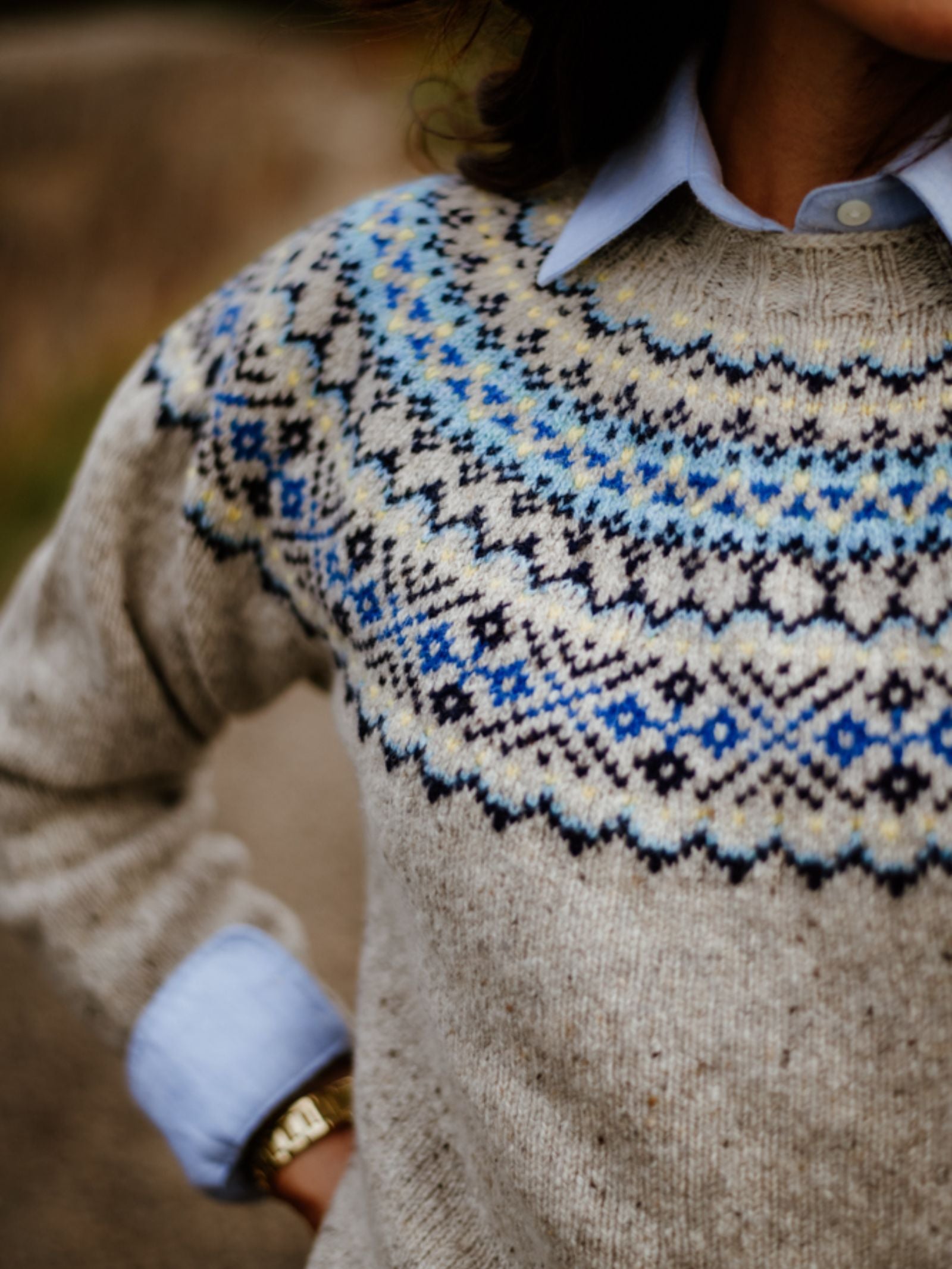 Someone stands with one hand on their hip, wearing a light blue collared shirt under the Campbell's of Beauly Donegal Lambswool Fairisle Crew Jumper in beige with a blue, black, and white Nordic pattern. Their head is cropped out of the frame.
