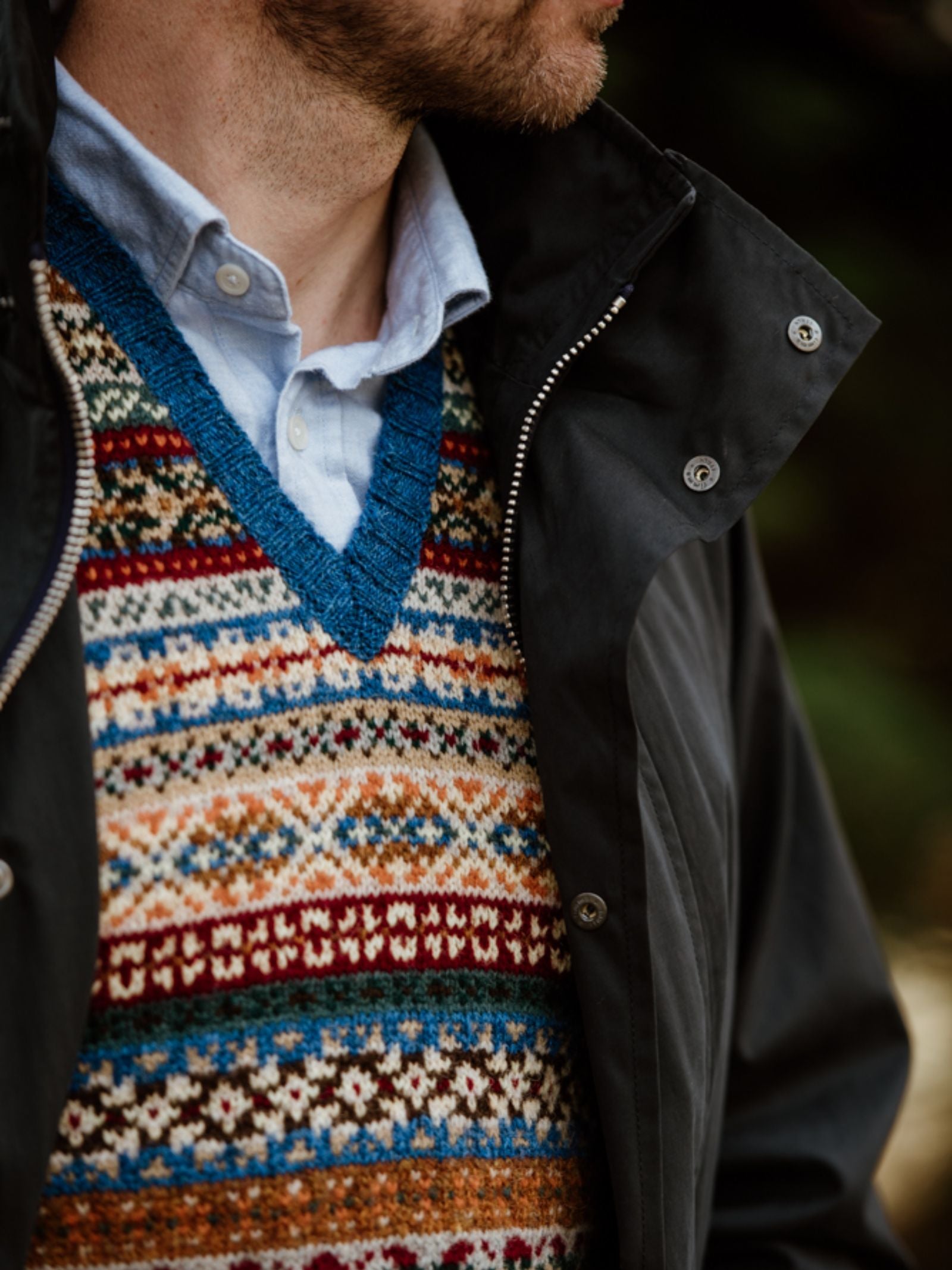 A man wears a blue collared shirt, a dark zippered jacket, and the Campbells of Beauly Fairisle Slipover—a sleeveless Shetland wool knit vest with blue, red, white, and gold patterns. The cropped photo shows only his lower face and torso.