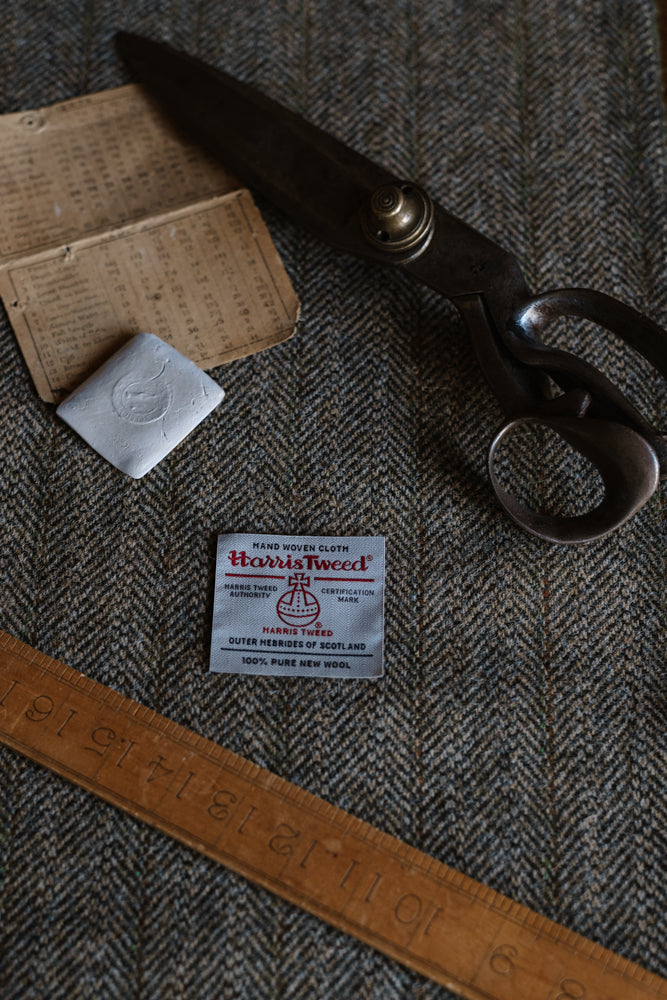 A House Tweed 95054 label from Campbell’s of Beauly rests on herringbone tweed fabric, accompanied by vintage scissors, a metal square, an old fabric chart, and a wooden ruler arranged on the tweed surface.