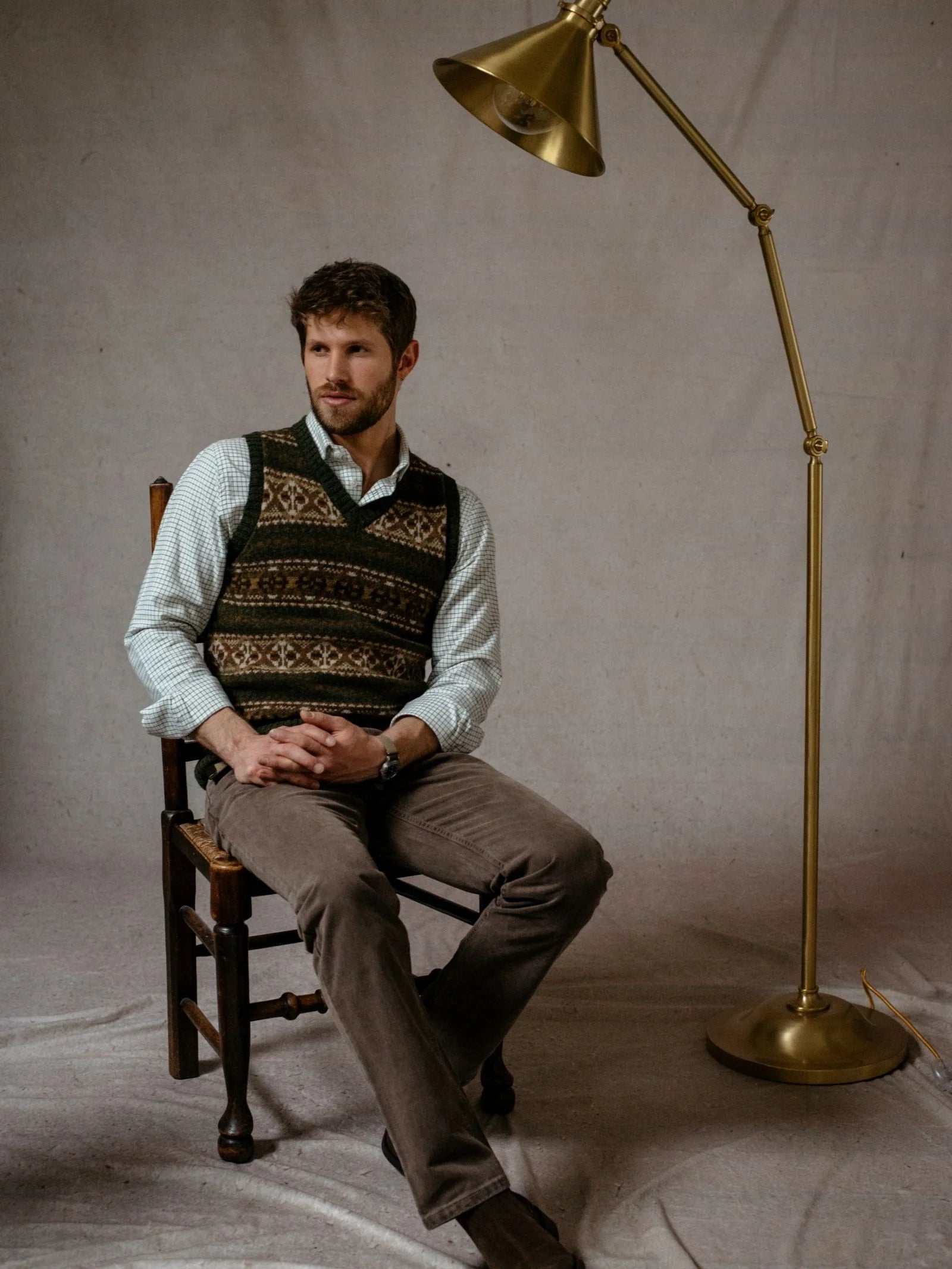 A man wearing the Campbell’s of Beauly Fairisle Slipover—sleeveless knitwear crafted from genuine Shetland wool—sits on a wooden chair beside a brass floor lamp, set against a plain backdrop.