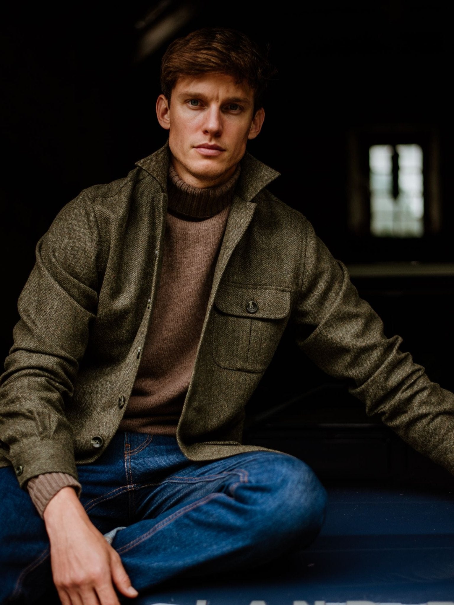 A young man with short brown hair sits casually on a dark surface, wearing blue jeans and a taupe sweater layered with the Campbell's of Beauly Casual Shacket. He gazes at the camera with a neutral expression, embodying relaxed tailoring.