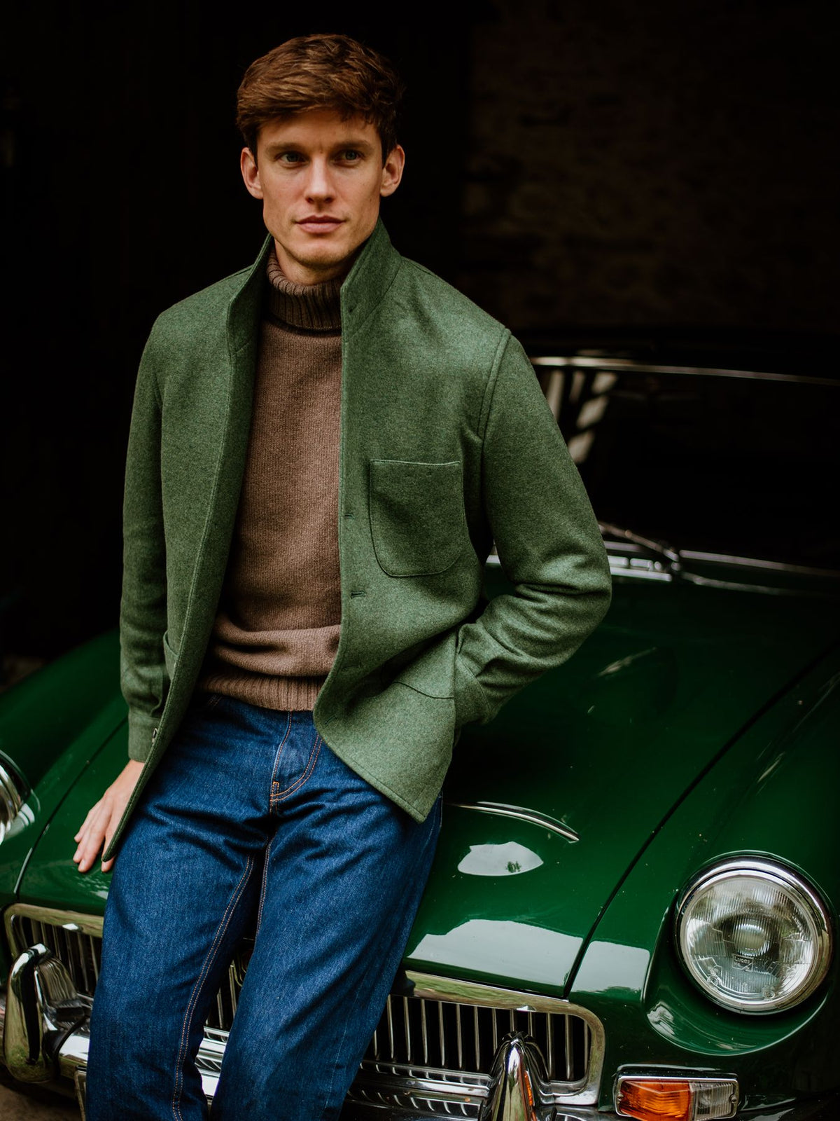 A man wearing a green jacket over a brown polo neck and blue jeans, leaning against a green vintage car.