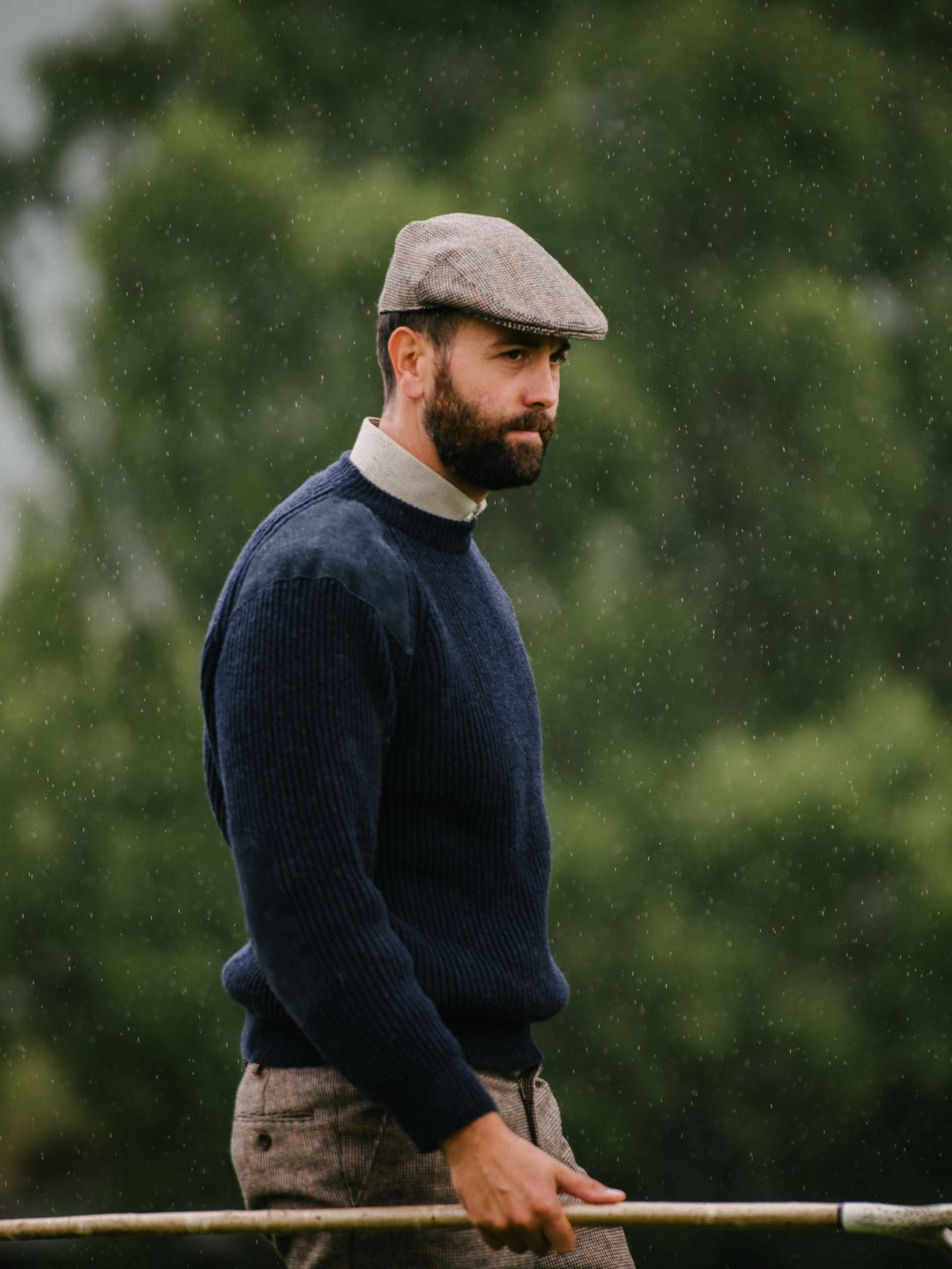 A bearded man in a Campbells of Beauly Scotia Suede Jumper, tweed trousers, and flat cap stands outdoors holding a wooden stick as rain falls around him, with blurred green trees suggesting a park or countryside backdrop.