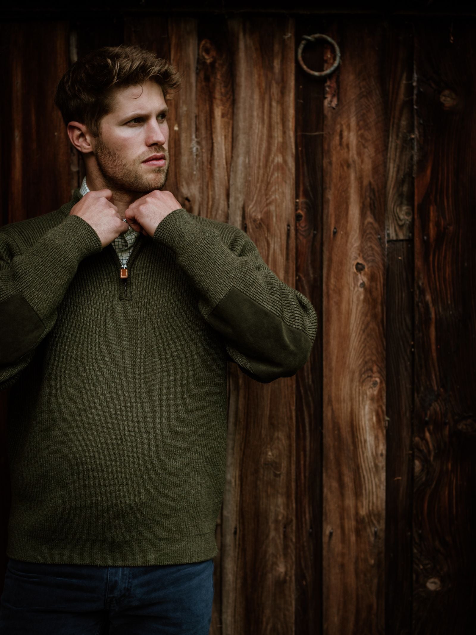 A man with short brown hair stands in front of a rustic wooden wall, thoughtfully adjusting the collar of his Campbells of Beauly Shooter Quarter Zip Jumper with Patches in forest green, paired with blue jeans.