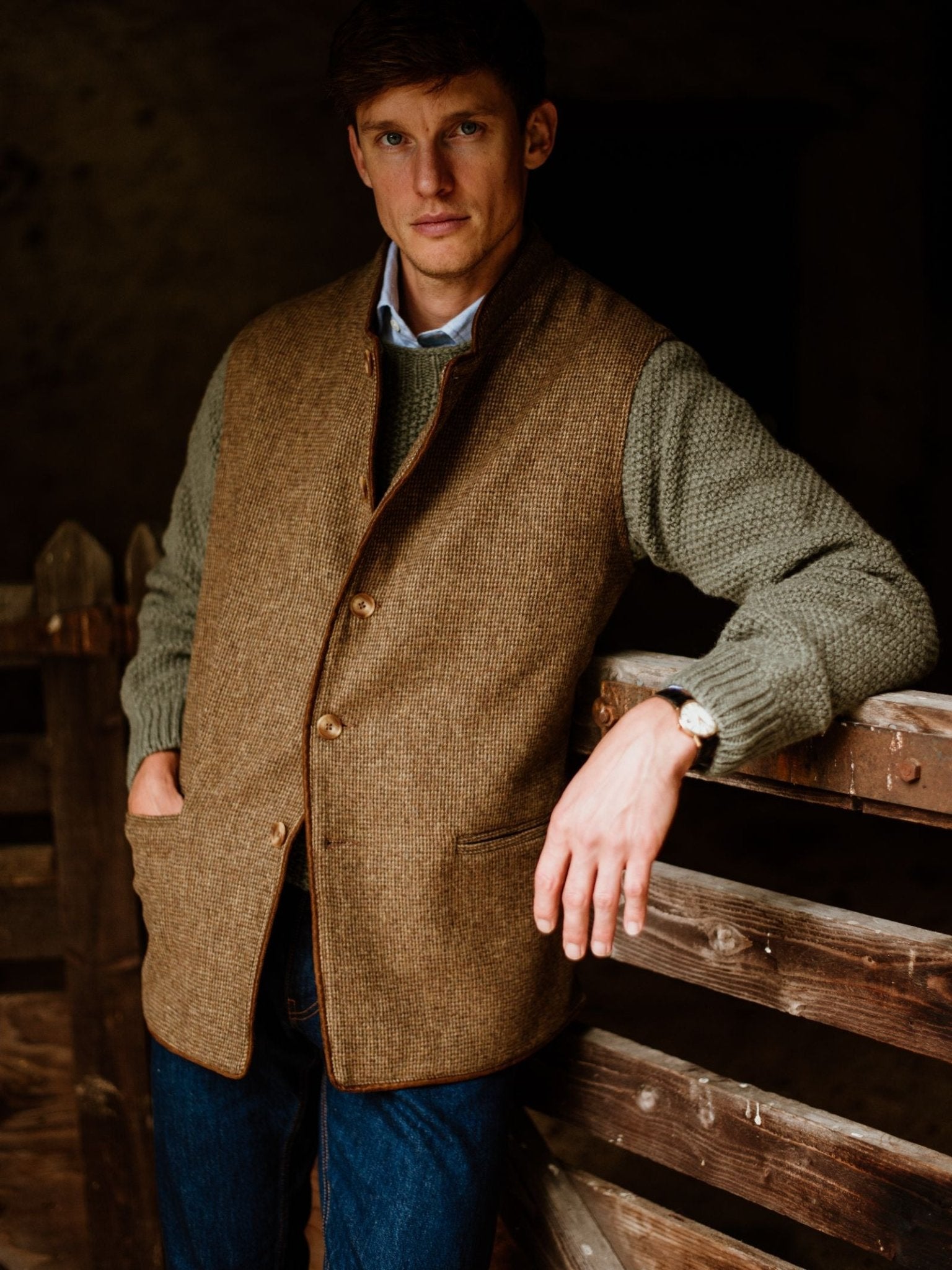A man wears the Campbell's of Beauly Tweed Nehru Gilet over a green sweater and blue jeans, leaning on a rustic wooden fence with a serious expression. The background is dark and blurred.