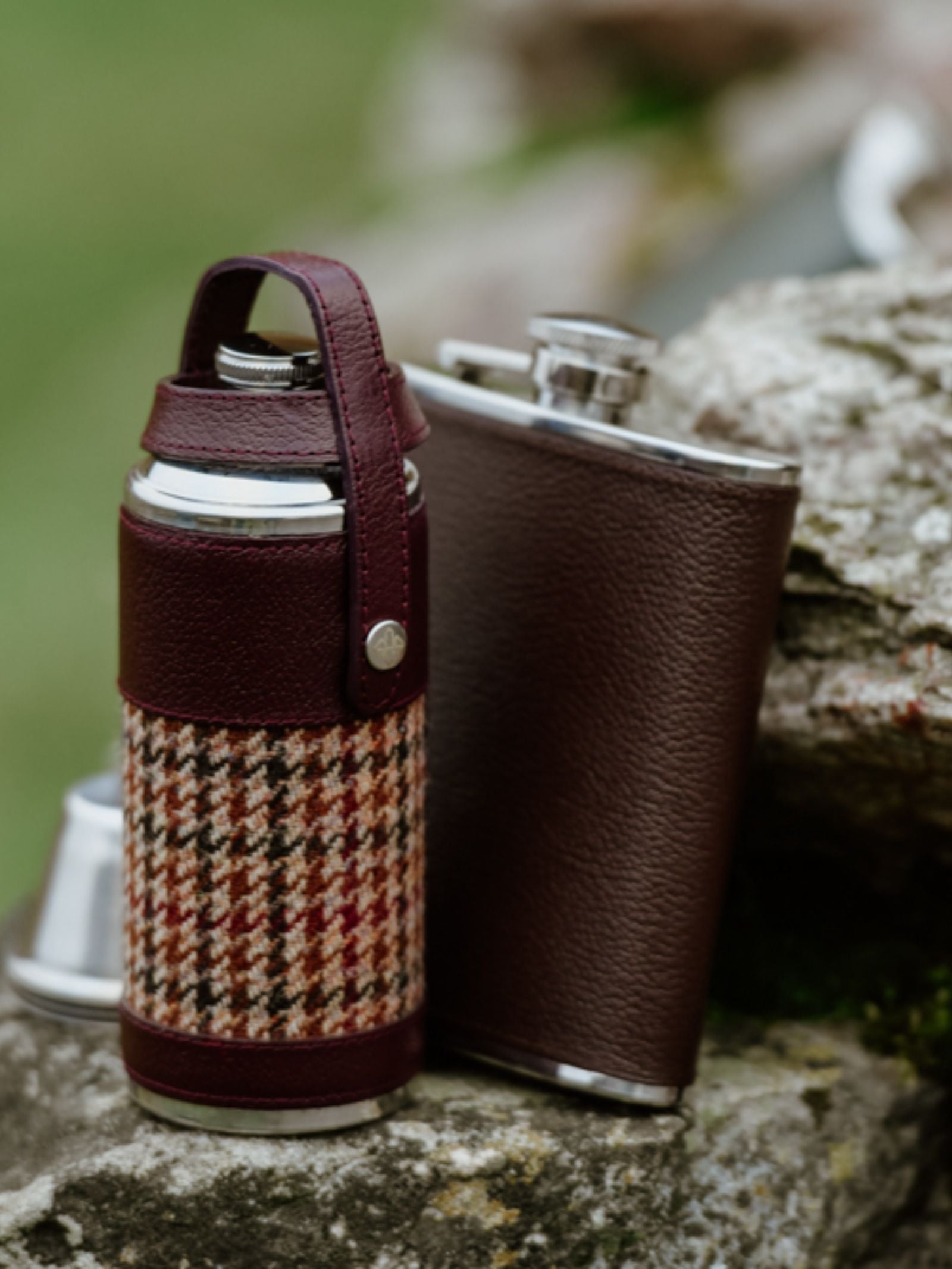 Two metal flasks rest on a rock outdoors. One is the Campbell's of Beauly x Ettinger 6oz Chocolate Hip Flask, wrapped in brown leather, while the other features Capra goat leather and patterned fabric. A small metal cup showcases British craftsmanship.