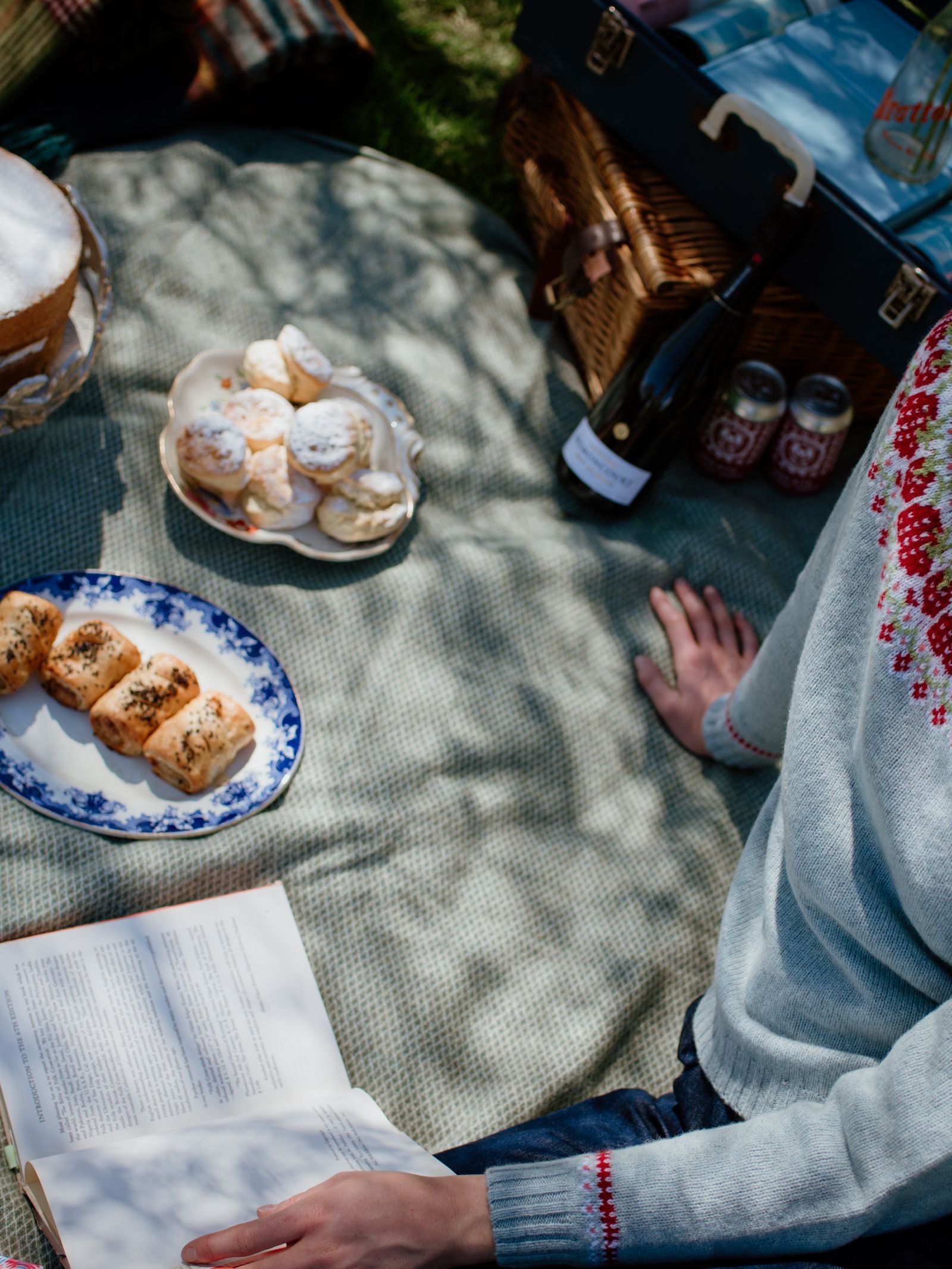 A person in a light blue sweater with red embroidery sits on a Campbells of Beauly Waterproof Picnic Blanket, holding an open book. Nearby are plates of pastries, a wine bottle, two jars, and a wicker basket as sunlight filters through the branches above.