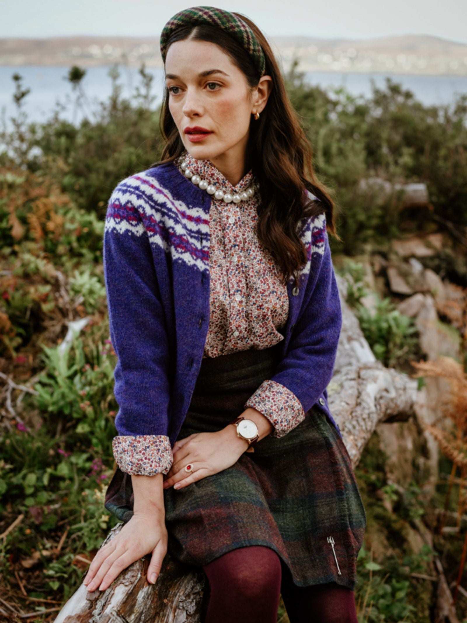 A woman with long dark hair sits outdoors on a log, surrounded by greenery and water, wearing a patterned headband, floral blouse, tartan skirt, maroon tights, and the Campbell's of Beauly Shetland Fairisle Cardigan in pure new wool.