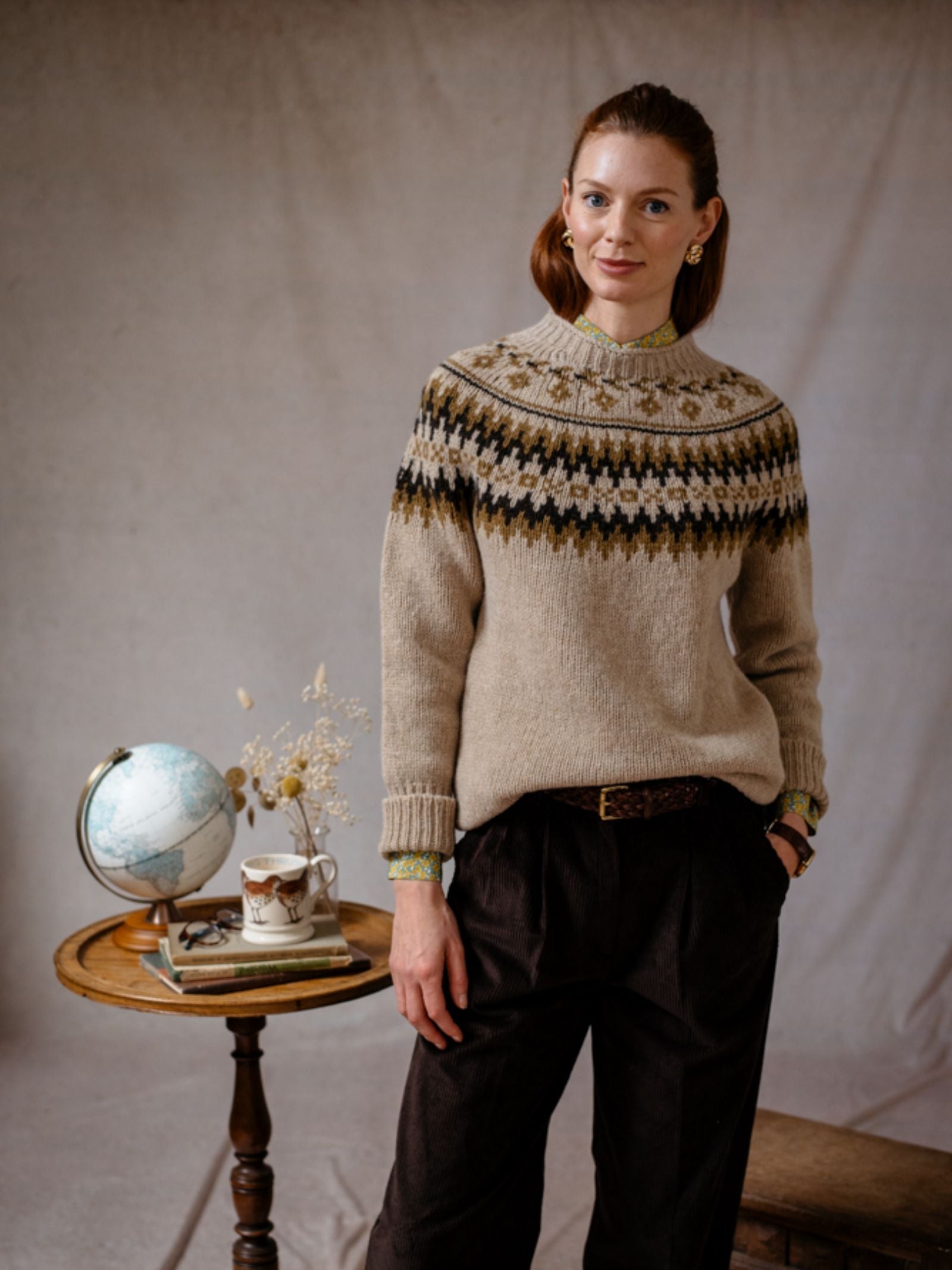 A light-skinned woman with brown hair wears the Chunky Fairisle Jumper by Campbells of Beauly and dark trousers, standing beside a small round table with a globe, books, dried flowers, and a teacup against a plain beige background. She smiles slightly, hands in pockets.