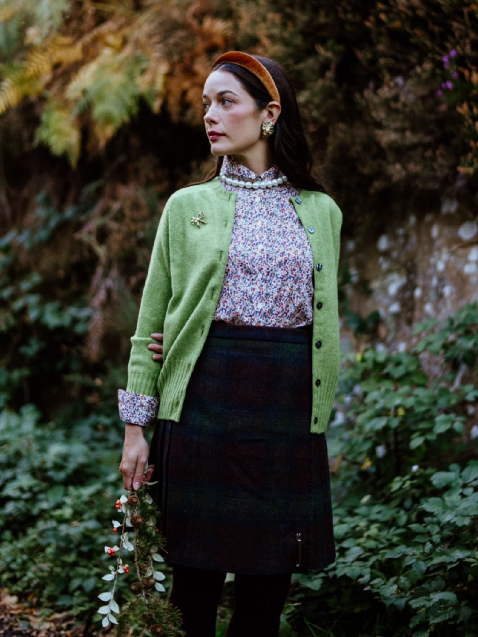 A woman stands outdoors among greenery, wearing Campbell's of Beauly Tweed Kilt with a green cardigan, floral blouse, and red headband. She holds a small bouquet of flowers and looks to her left.