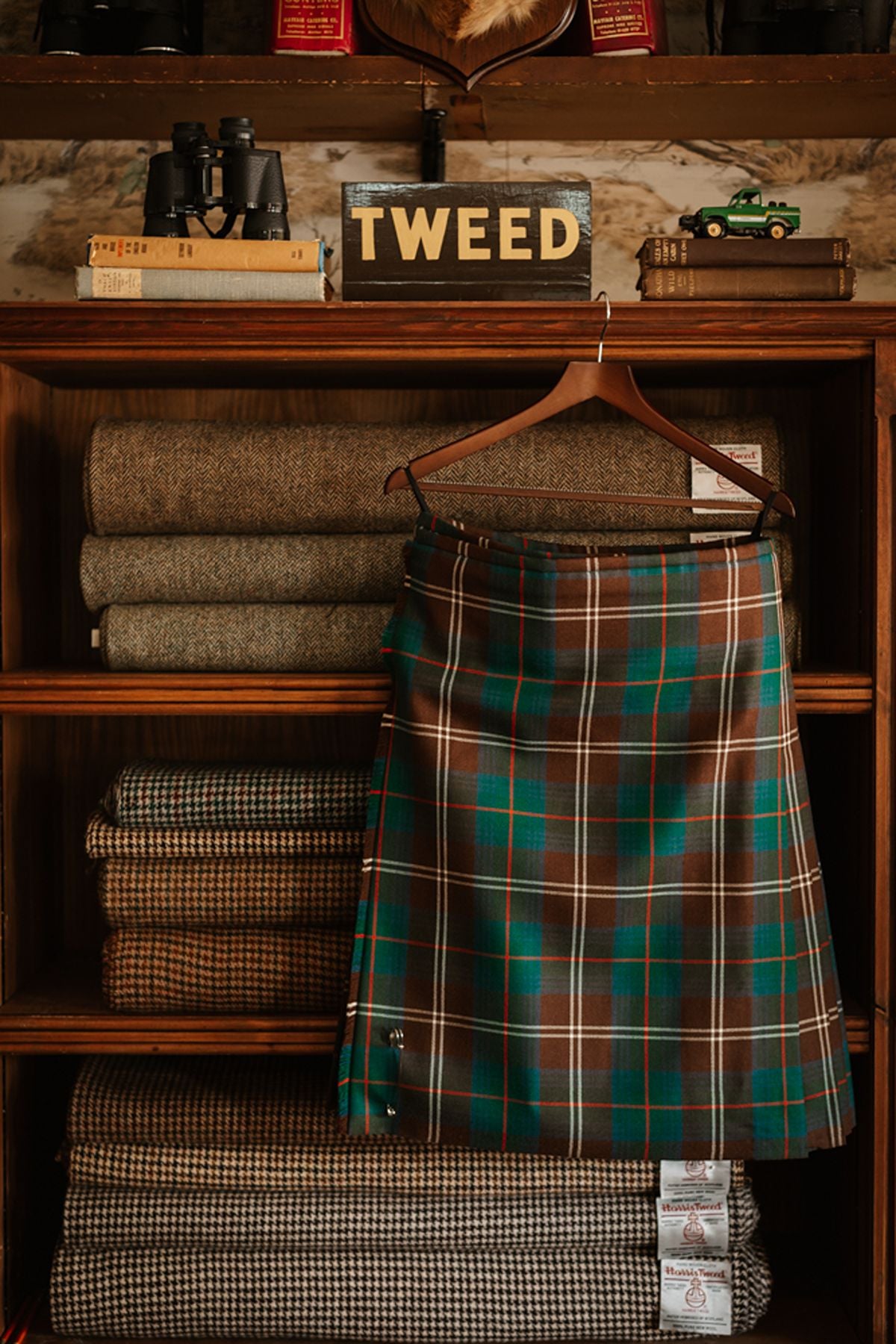 A green, brown, and white tartan kilt hangs in front of shelves stocked with folded rolls of tweed fabric.