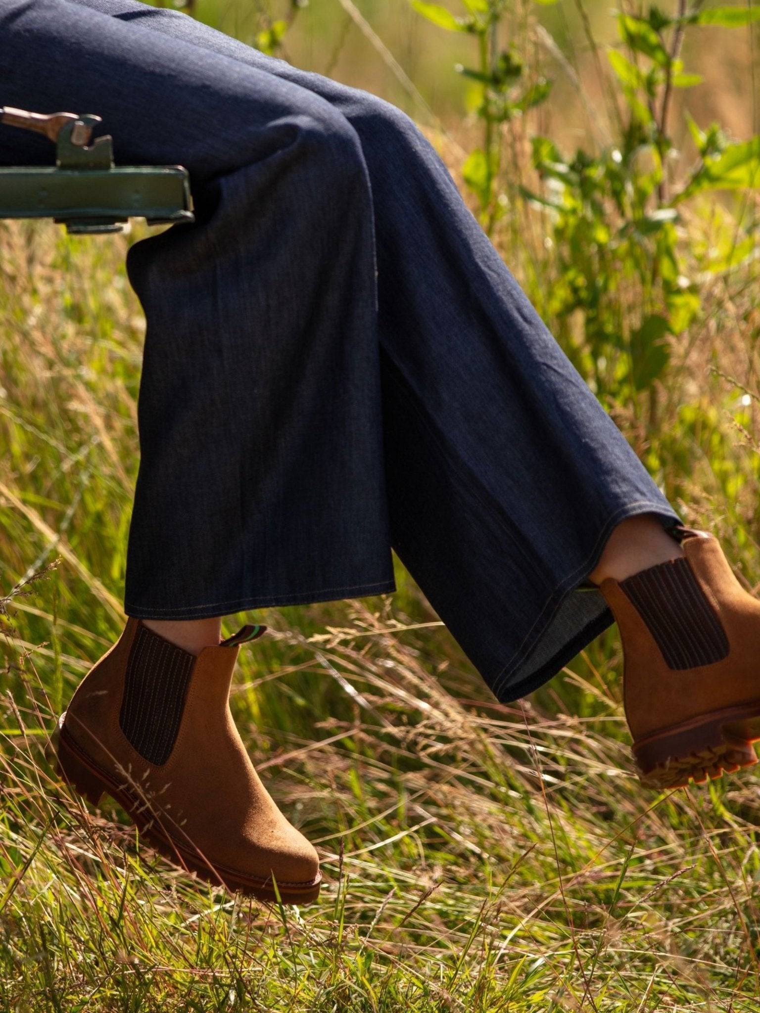 Wearing Campbell's of Beauly Leather Chelsea Boots and wide-leg blue jeans, a person sits on a green bench amid tall grass—capturing the perfect classic country wardrobe for an outdoor setting.