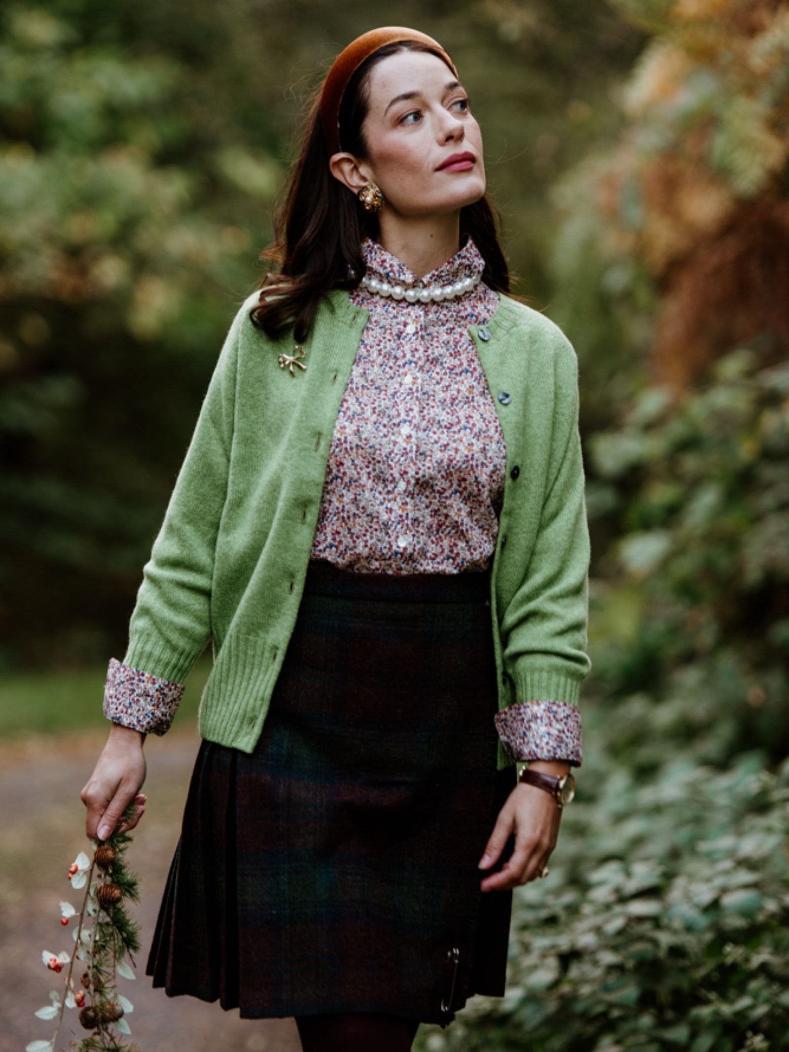 A woman stands outdoors on a leafy path, wearing the Campbell's of Beauly Crew Cardigan in green over a floral blouse and dark plaid skirt, with a brown headband. She holds flowers and gazes thoughtfully into the distance.
