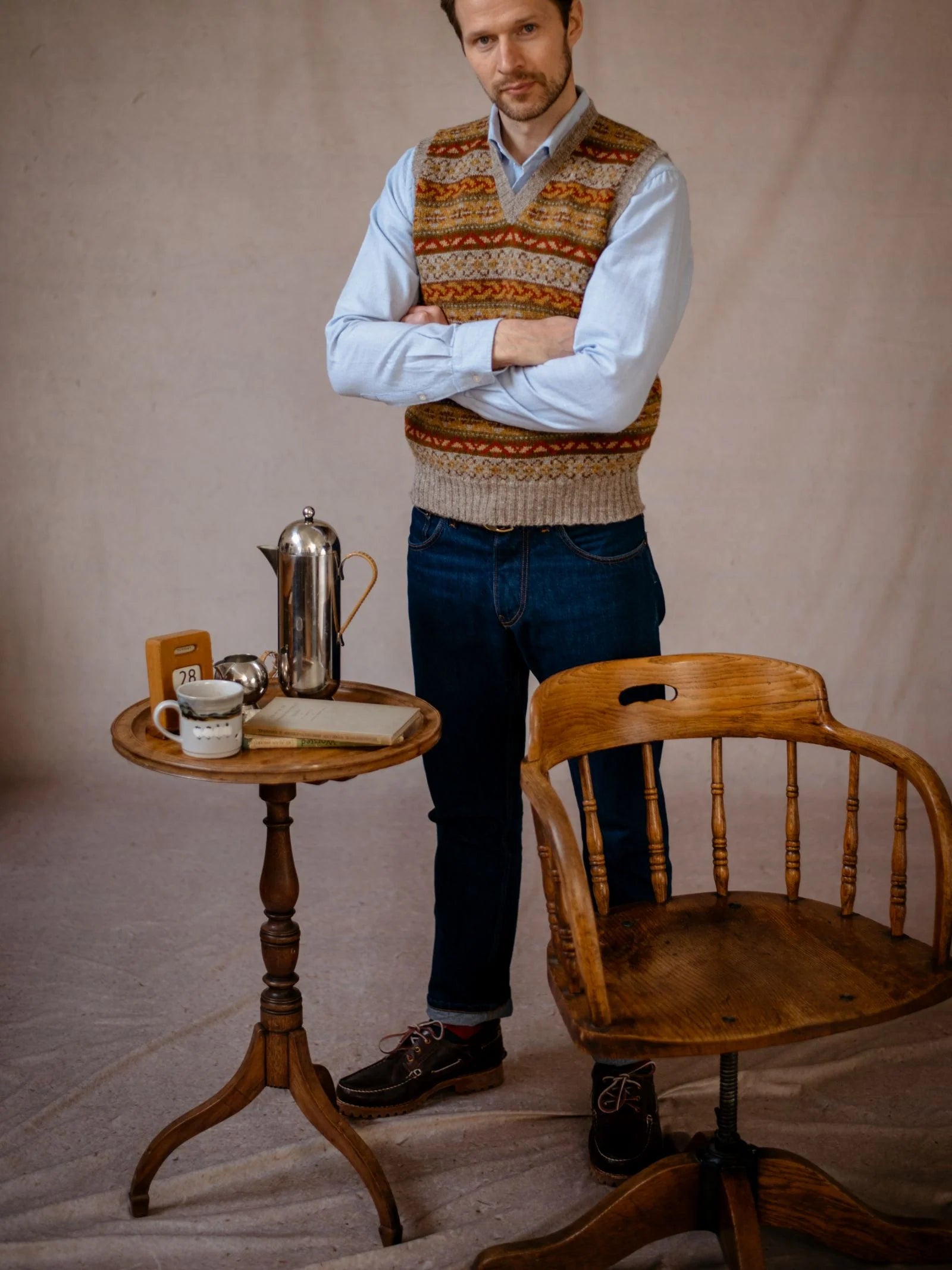 A man wearing the Campbell’s of Beauly Fairisle Slipover—a classic sleeveless layer knit from soft Shetland wool—paired with jeans, stands beside a small round wooden table set with a coffee pot, cup, and book. An empty wooden chair sits in front of him against a neutral background.