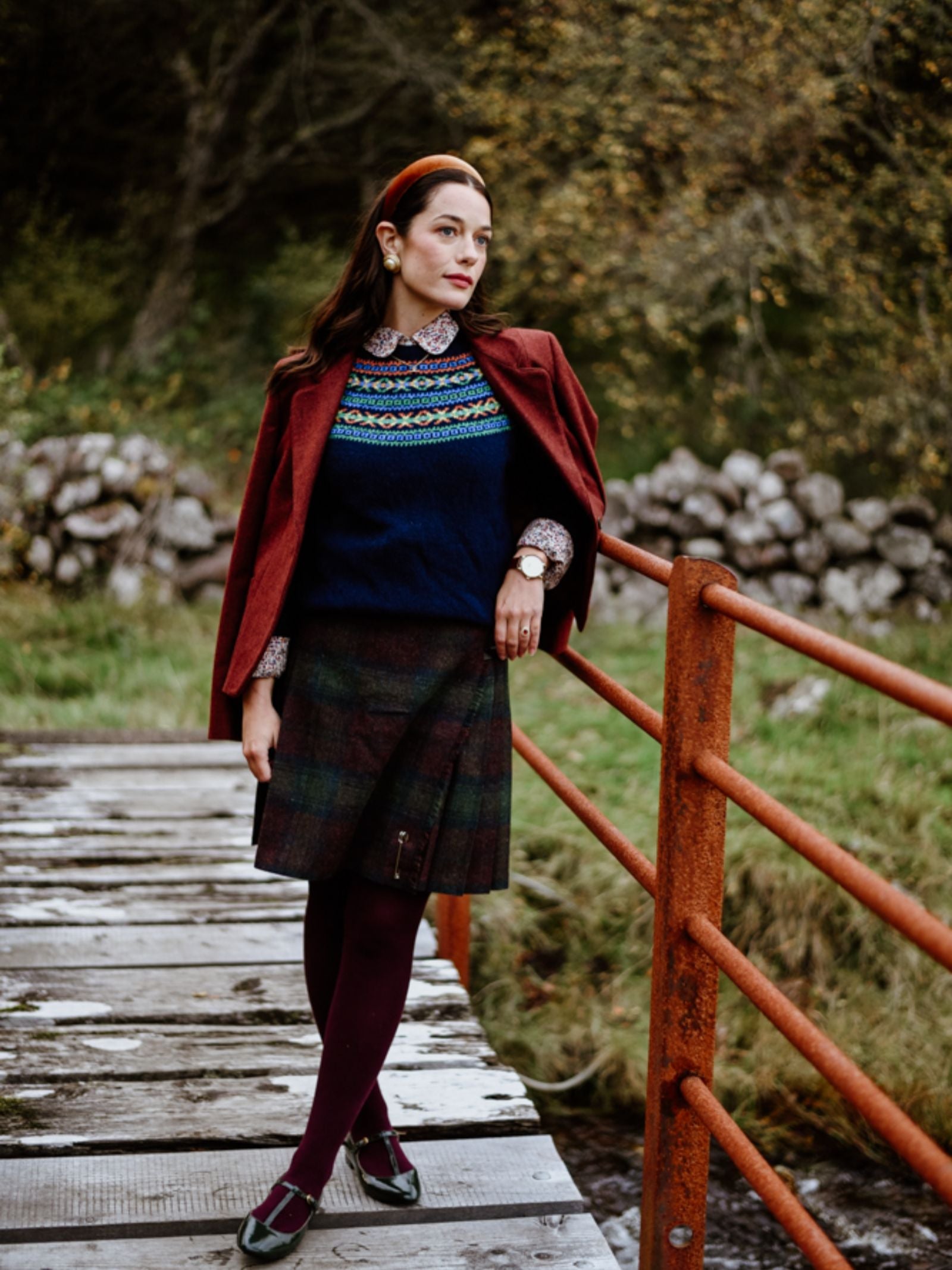 A woman stands on a wooden bridge wearing a red headband, maroon jacket, patterned sweater, Campbell's of Beauly Tweed Kilt, burgundy tights, and black shoes, with trees and rocks in the background.