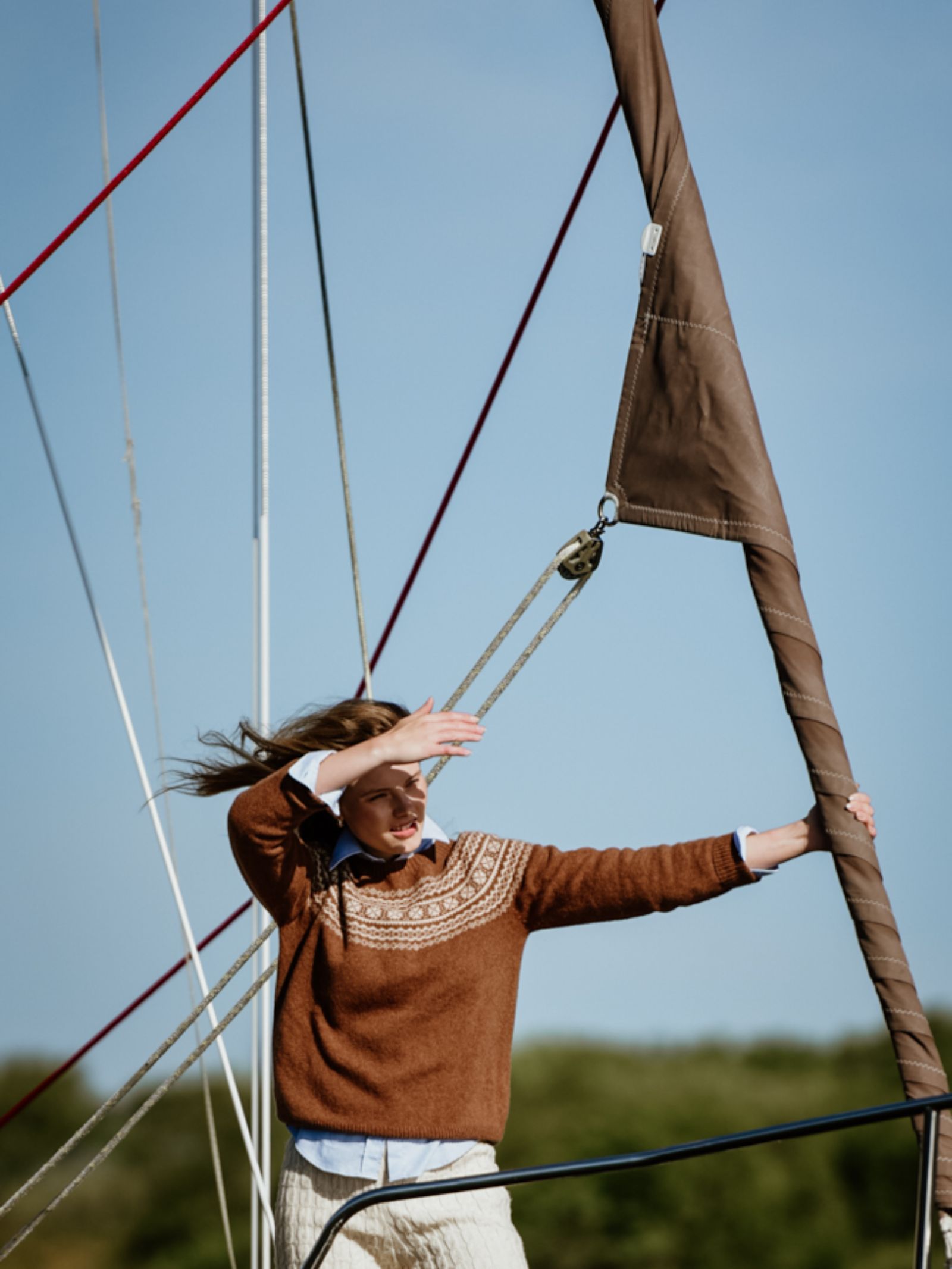 Wearing the Campbells of Beauly Two-Colour Fairisle Yoke Jumper in superfine Geelong lambswool, a person stands on a sailboat, shielding their eyes. A clear sky and blurred trees evoke the spirit of Scottish craftsmanship.