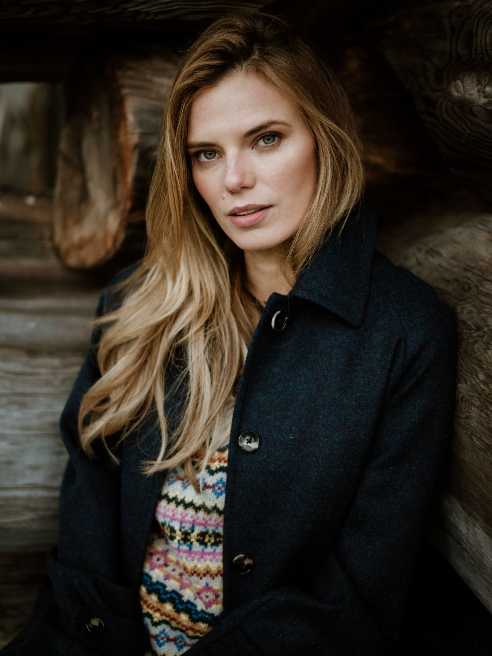 A woman with long blonde hair, wearing the Campbells of Beauly Midnight Collar Coat and a colorful patterned sweater, sits calmly in front of stacked wooden logs, looking at the camera.