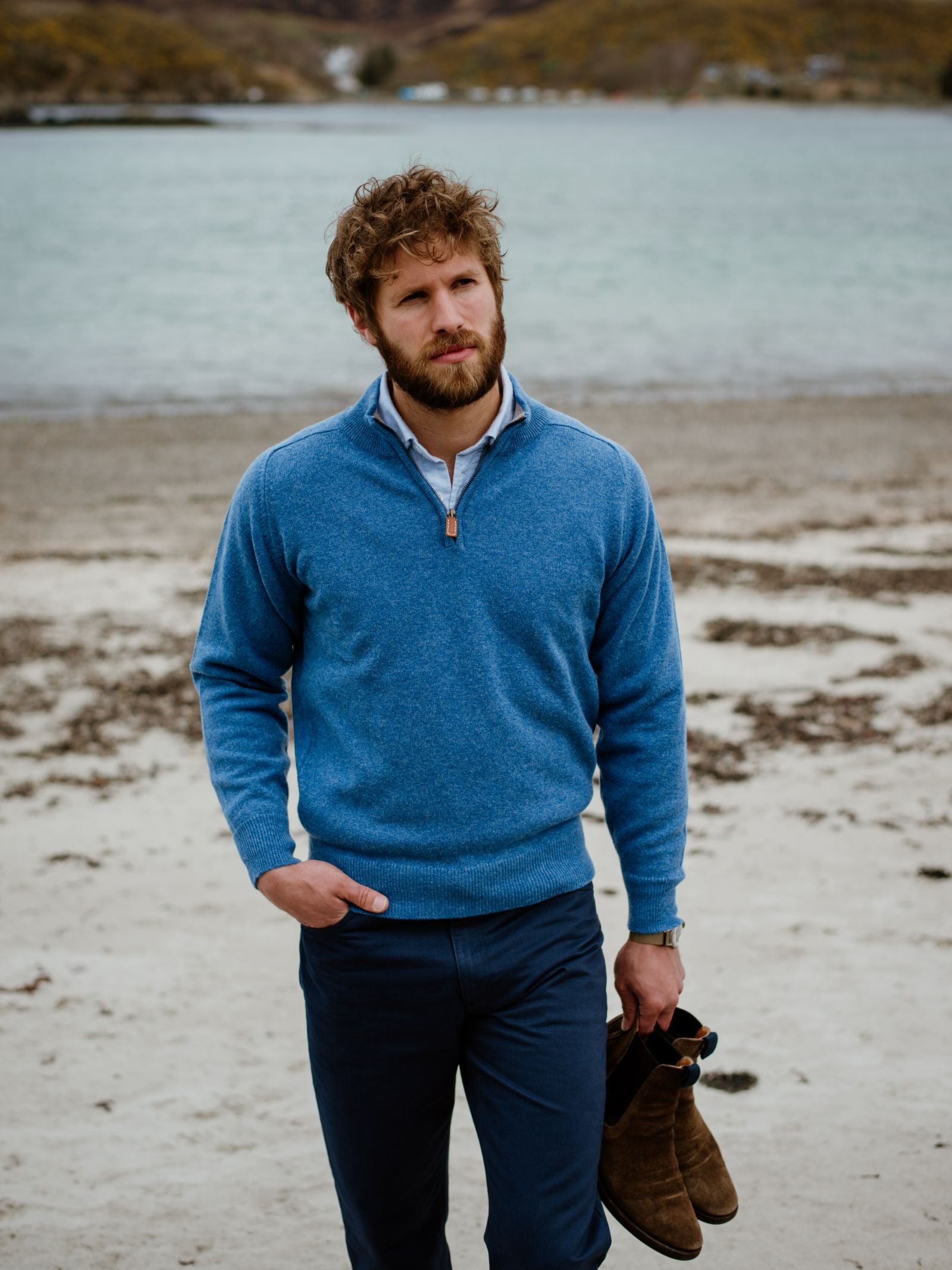 A man with curly hair and a beard walks on a sandy beach in the Scottish Borders, wearing the Campbells of Beauly Lambswool Quarter Zip Jumper in blue and dark pants, holding brown boots as he gazes thoughtfully at the calm water and distant hills.