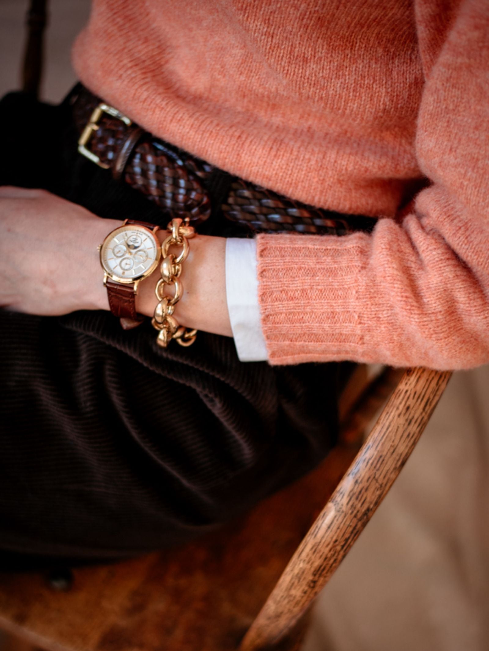 Seated on a wooden chair, the person wears Campbells of Beauly Cropped Shetland Jumper in peach lambswool over a white cuff, paired with dark corduroy pants, a braided brown belt, and accessorizes with gold jewelry and a brown leather-strap watch.