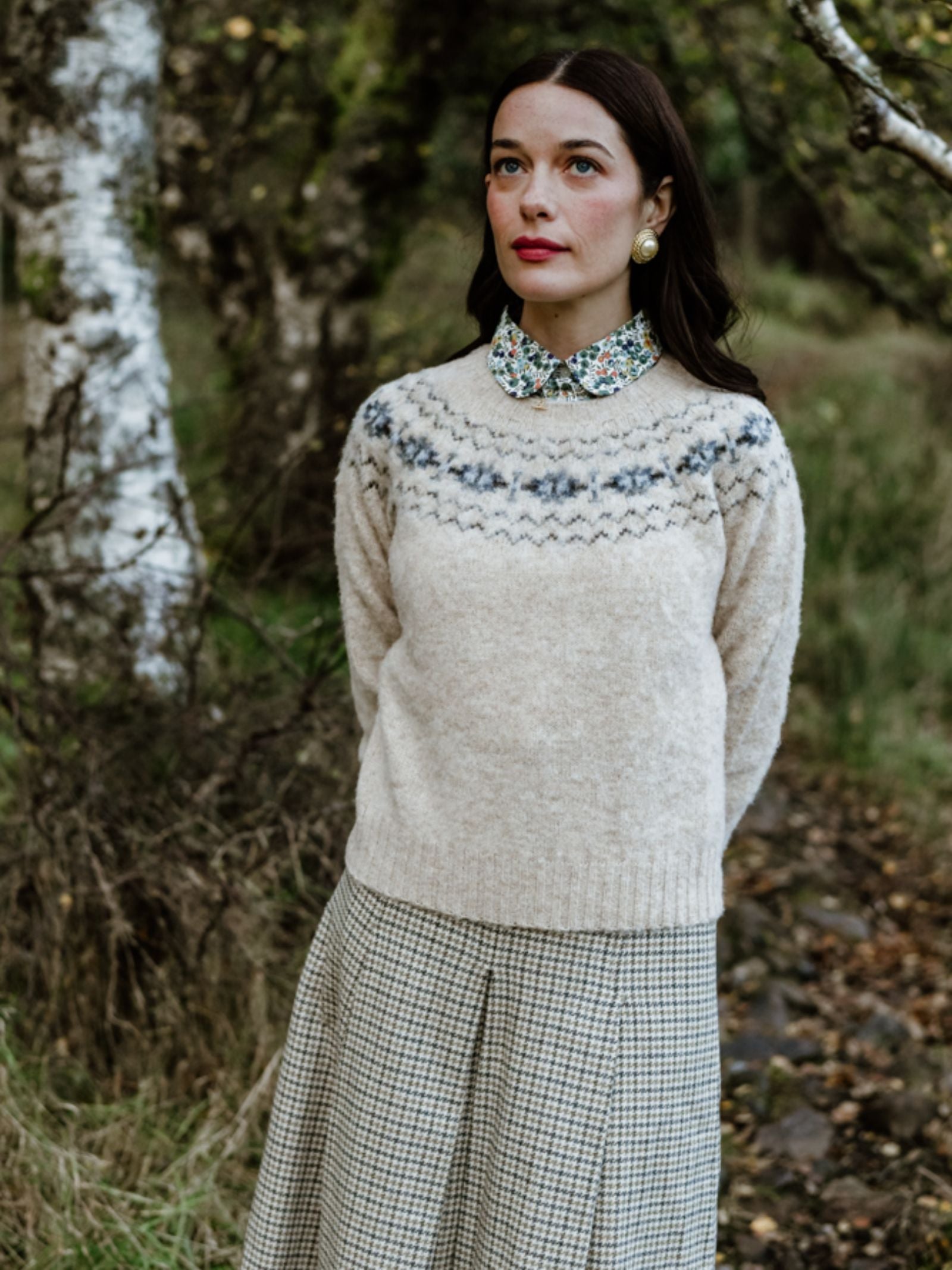 A woman with long brown hair stands outdoors by trees, wearing a Campbell's of Beauly Cropped Fairisle Crew Jumper with a patterned yoke over a floral shirt, paired with a plaid skirt and large round earrings, gazing slightly upward.