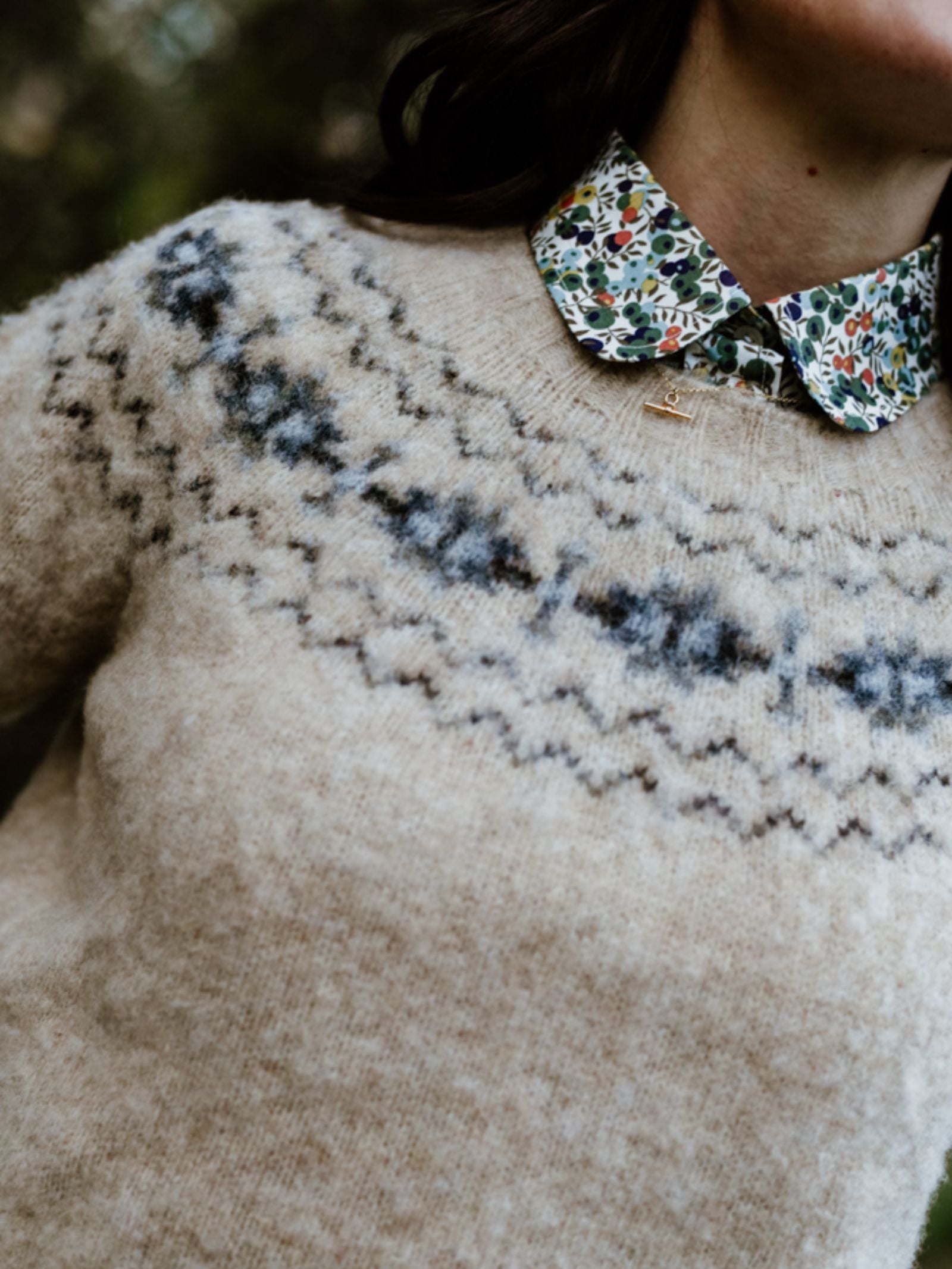 A person wears the Campbell's of Beauly Cropped Fairisle Crew Jumper in beige with a blue zigzag pattern, layered over a floral collared shirt. The outdoor photo highlights the cropped fit and focuses on the upper chest and neck.