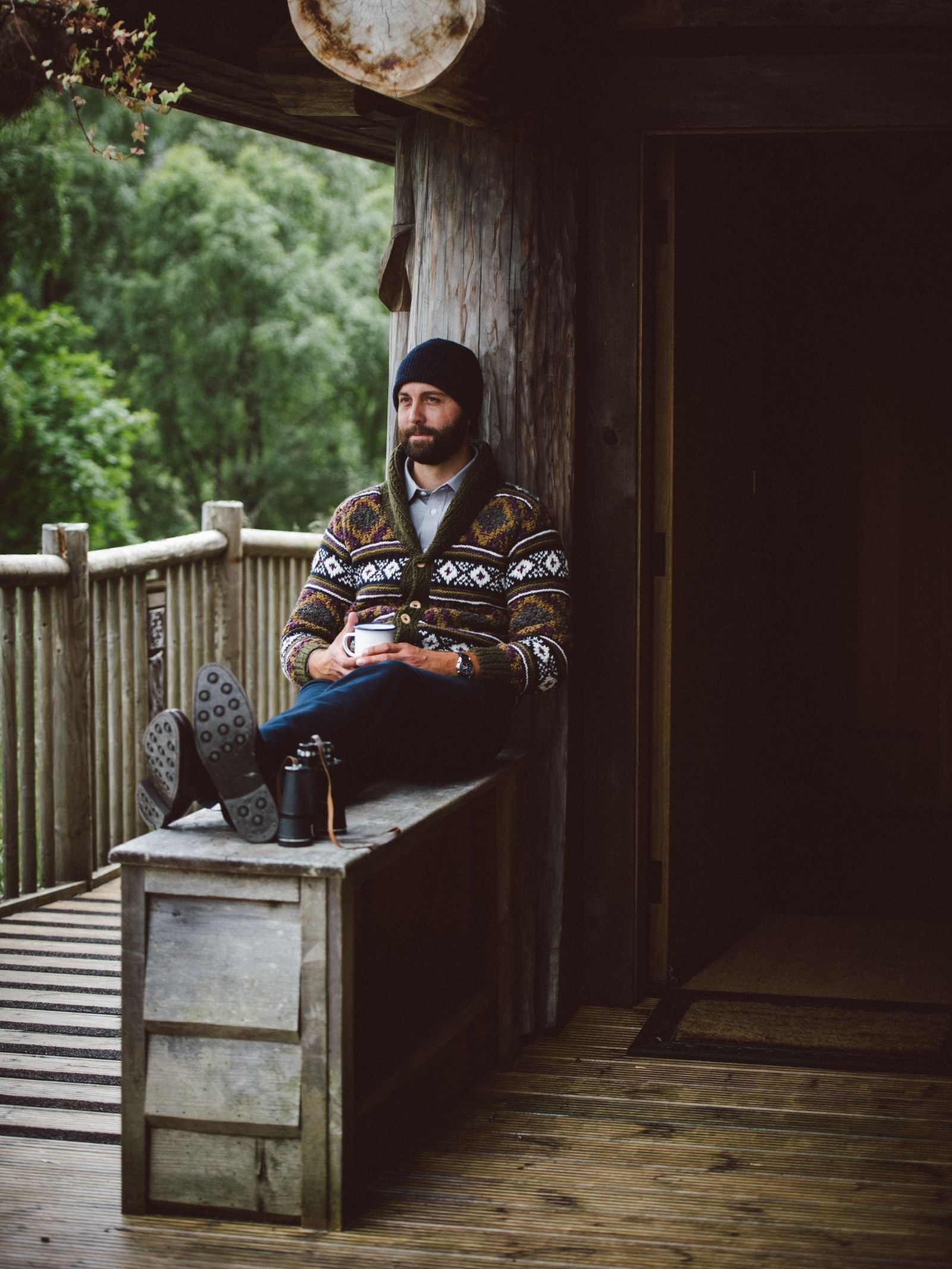 A bearded man in a Campbells of Beauly Navajo Shawl Collar Cardigan sits on a rustic wooden chest on a porch, holding a mug. He looks relaxed beside his camera, enjoying the cozy, cabin-like atmosphere and forest view.