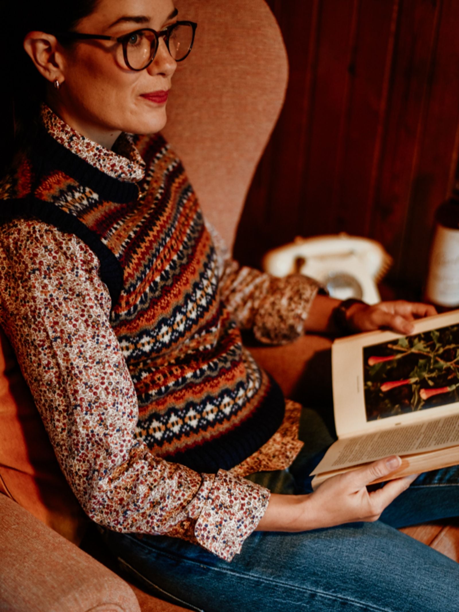 A person wearing glasses, a Campbell's of Beauly Fairisle Crew Tank and a floral shirt sits in an armchair, reading a botanical book in a cozy, warmly-lit room.