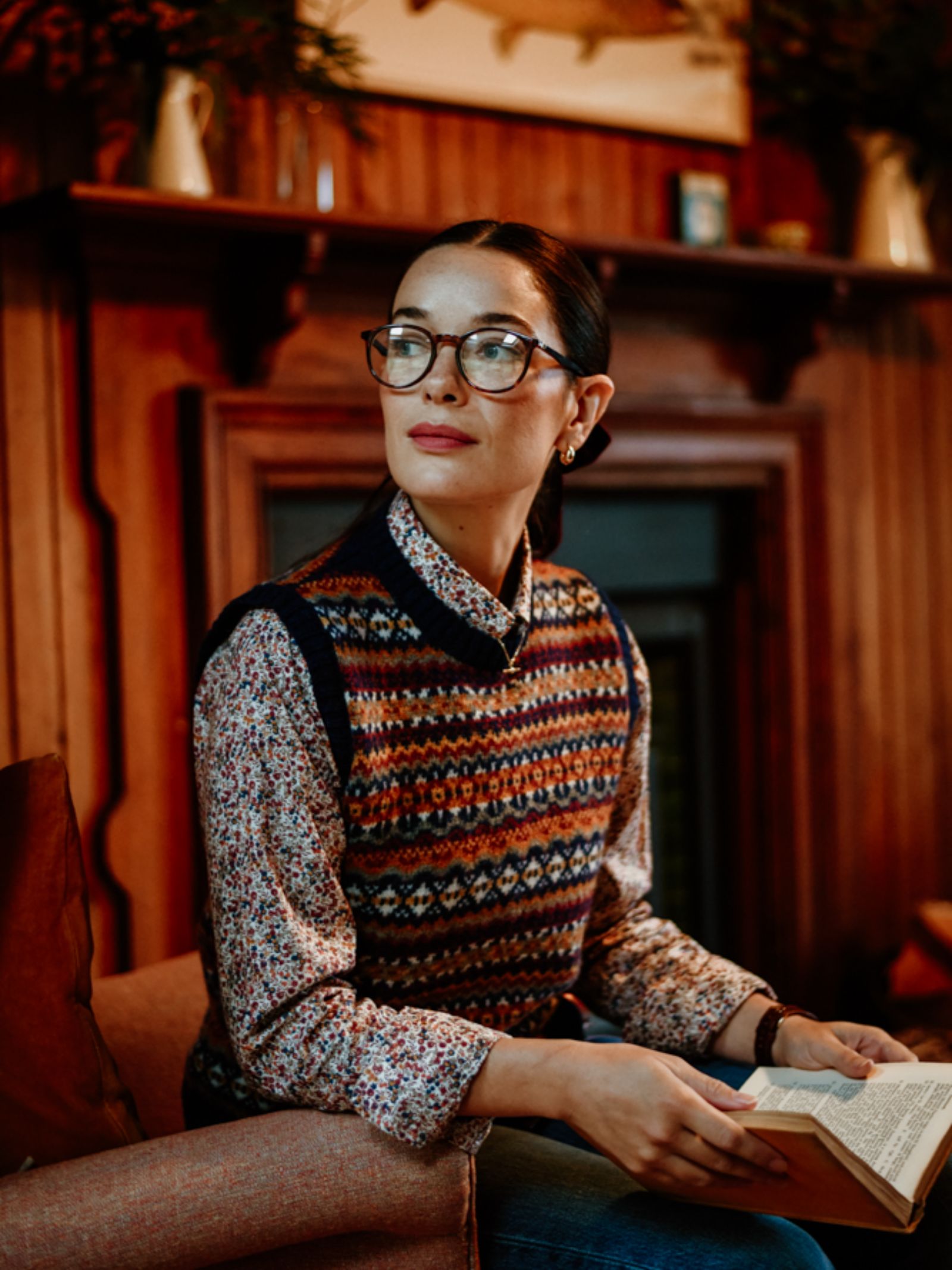 A woman in glasses wears the Campbell's of Beauly Fairisle Crew Tank over a floral shirt, sitting on a couch with an open book in a cozy, wood-paneled room, a fireplace glowing softly in the background.